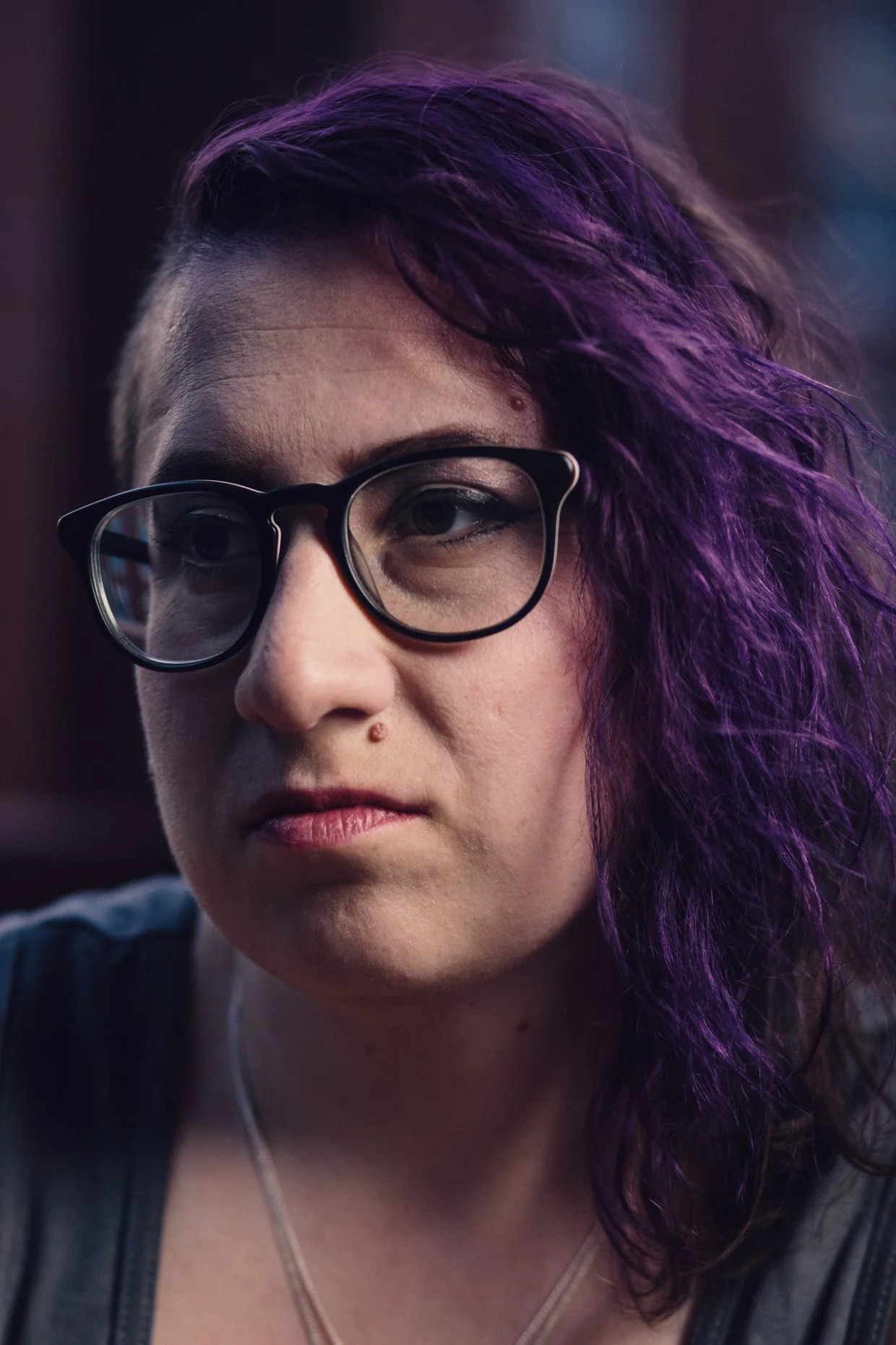 Close-up of a woman with purple curly hair, wearing black glasses and a serious expression.