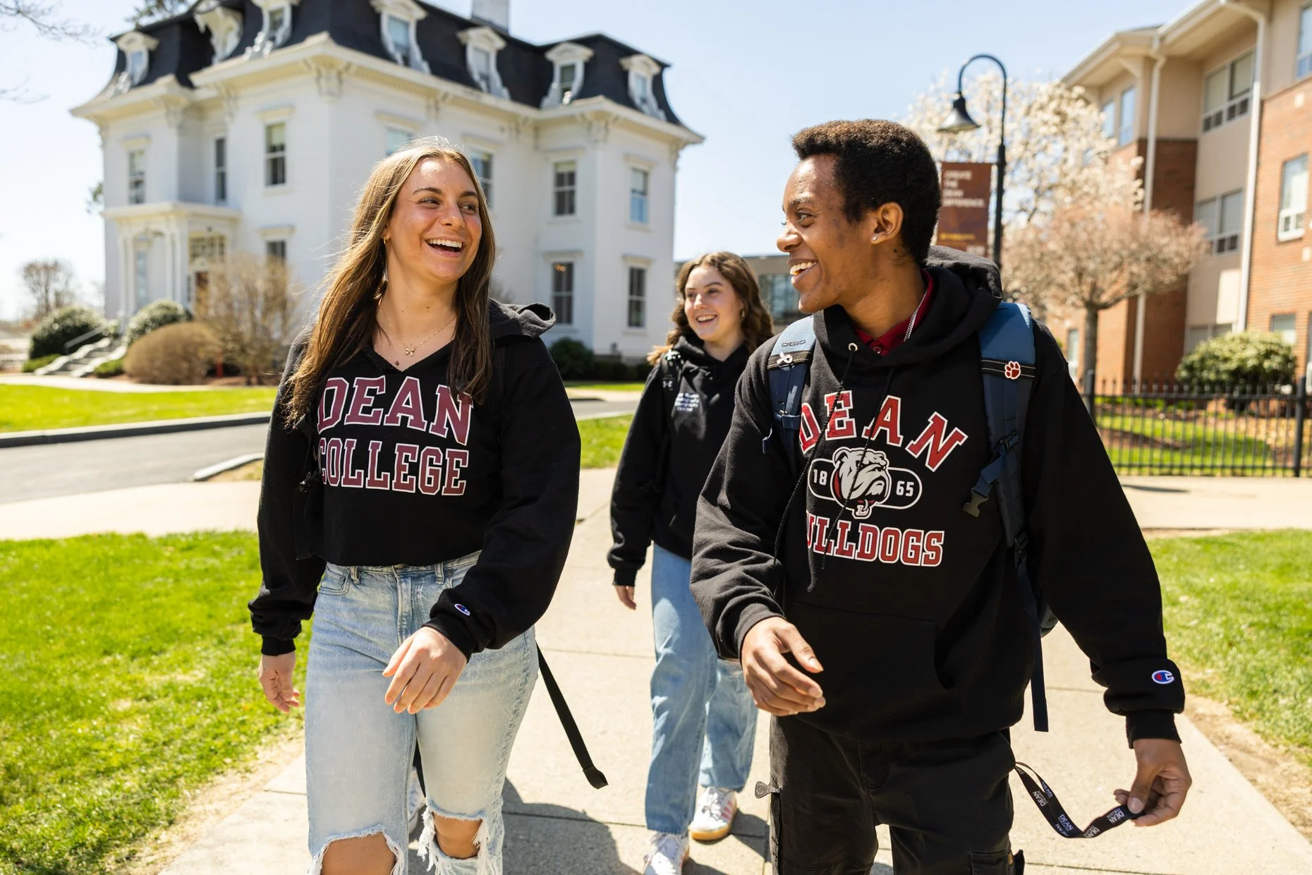Three college students walking outside on a sunny day, smiling and laughing, wearing black sweatshirts with 'Dean Bulldogs' printed on them, with a white house and other campus buildings in the background.