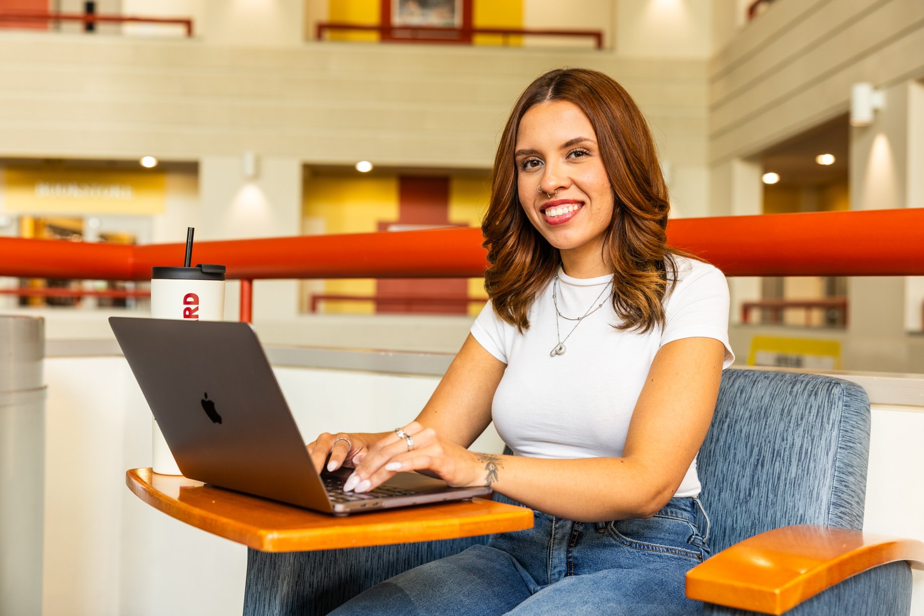 A young woman with reddish-brown hair, wearing a white t-shirt and jeans, sitting at a wooden table in an indoor public space. She is smiling at the camera while typing on a laptop, with a white travel tumbler featuring red text next to her.