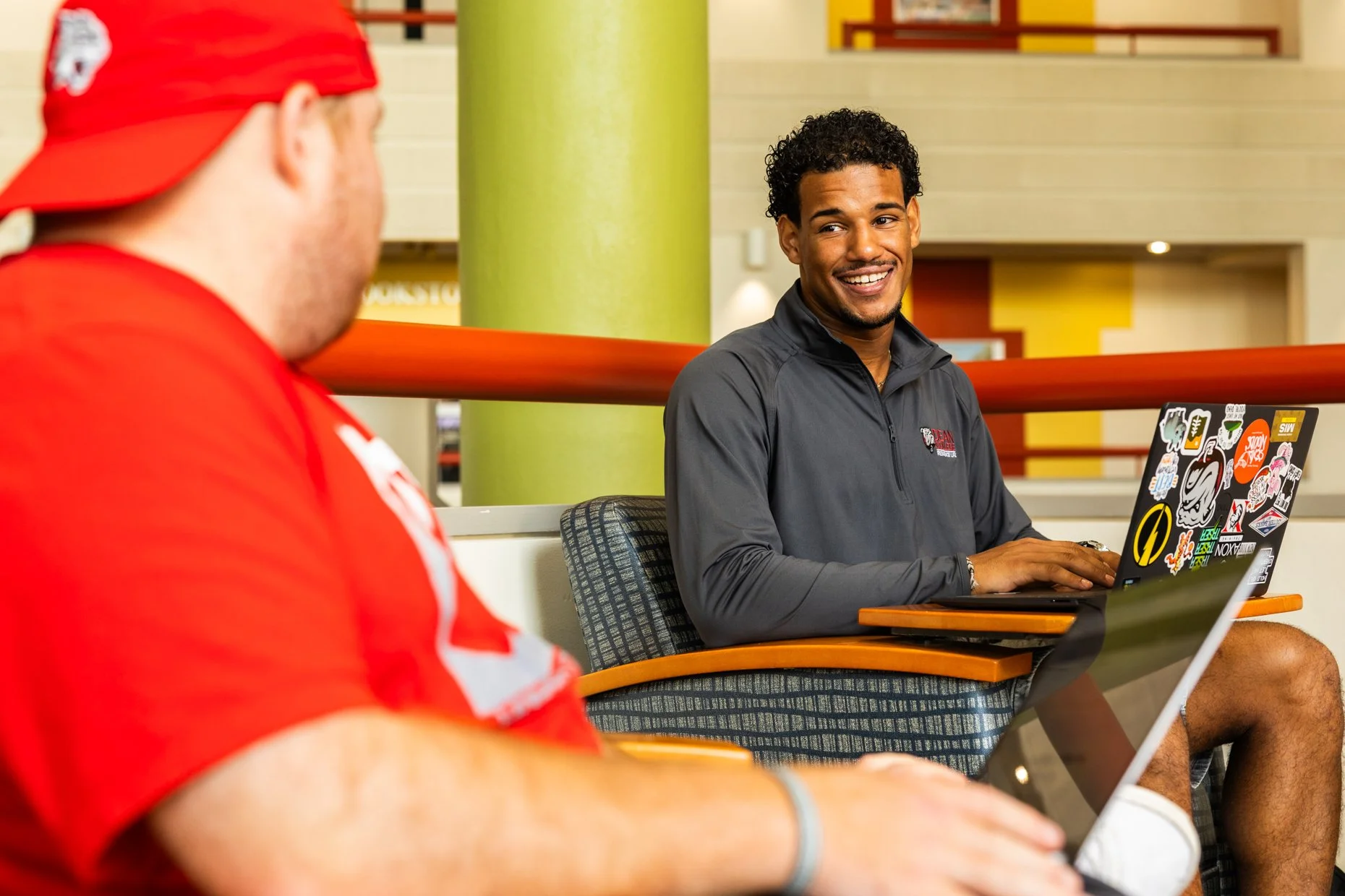 Two men sitting at a table in a modern indoor space, with one man wearing a red cap and red shirt, and the other man in a gray long-sleeve shirt with a laptop covered in stickers, smiling and engaged in conversation.
