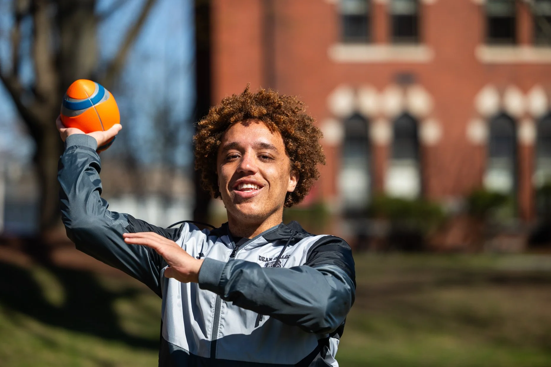 A young man with curly hair is outdoors, holding a colorful basketball on his shoulder while smiling. He is wearing a black, white, and gray athletic jacket, and the background includes blurred trees and a brick building.