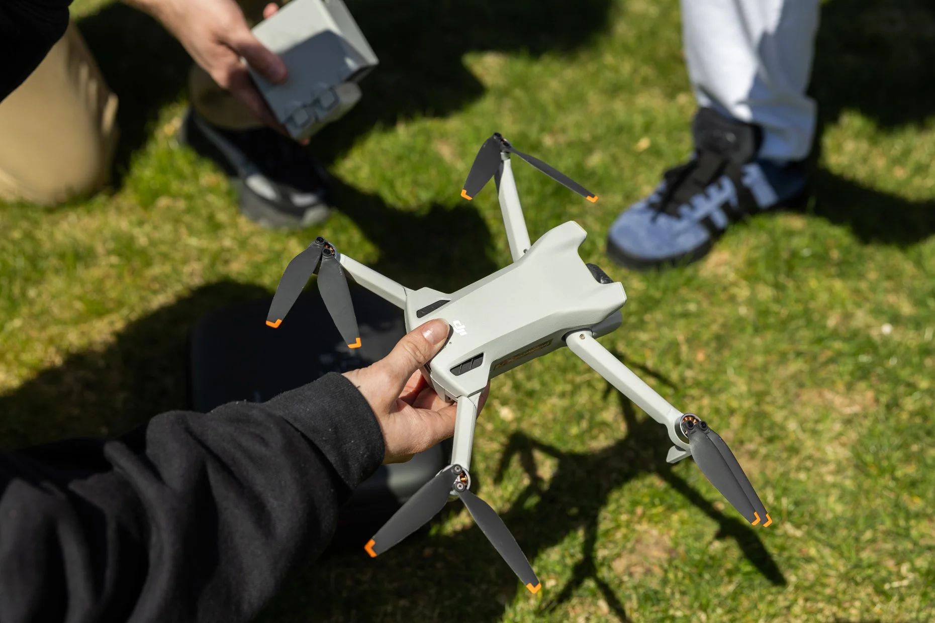 Person holding a DJI drone with three blades each marked with orange tips, positioned outdoors on green grass, with two other people nearby.