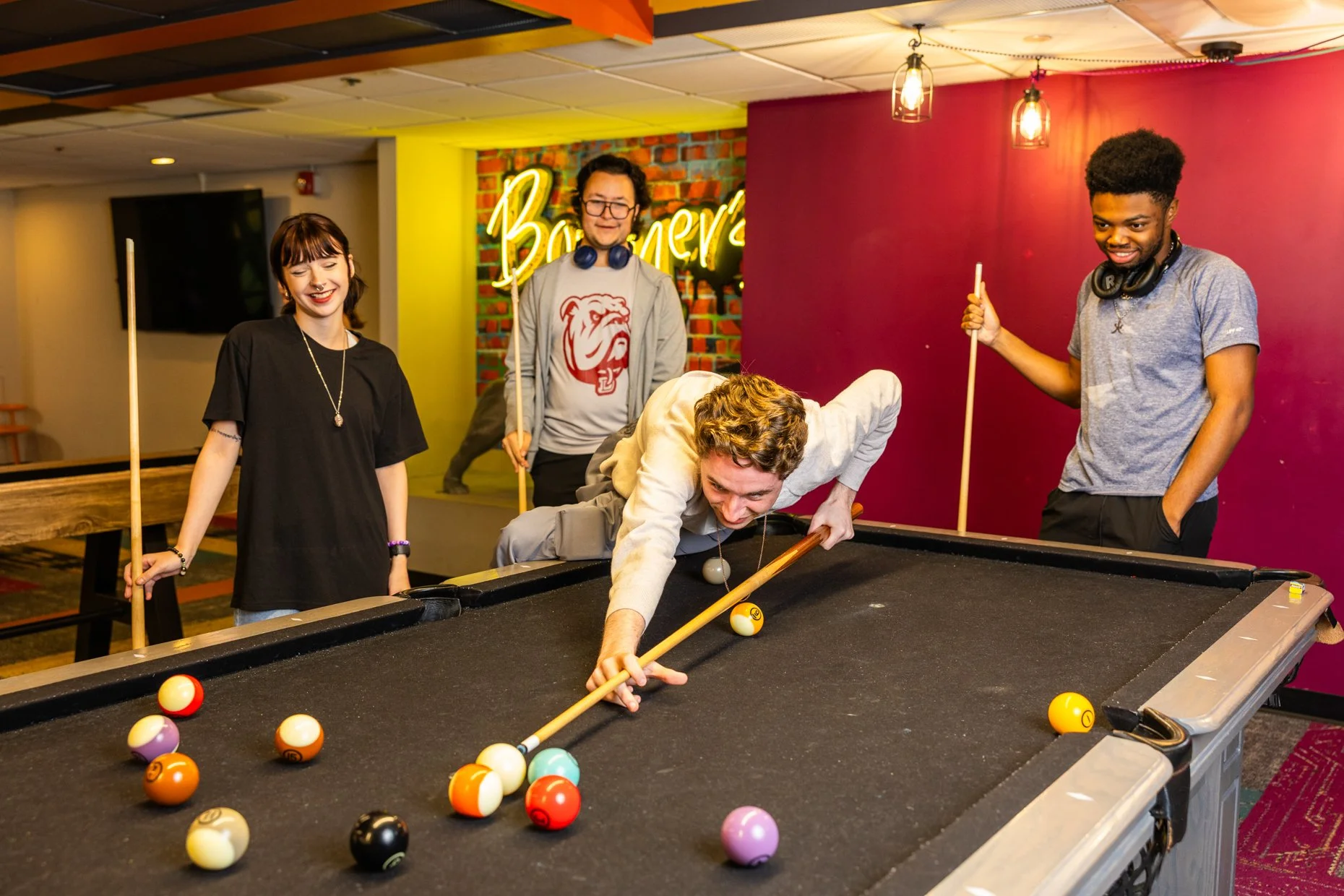 Four young adults playing pool in a casual entertainment space, with a colorful background and neon sign reading 'Bowery'.