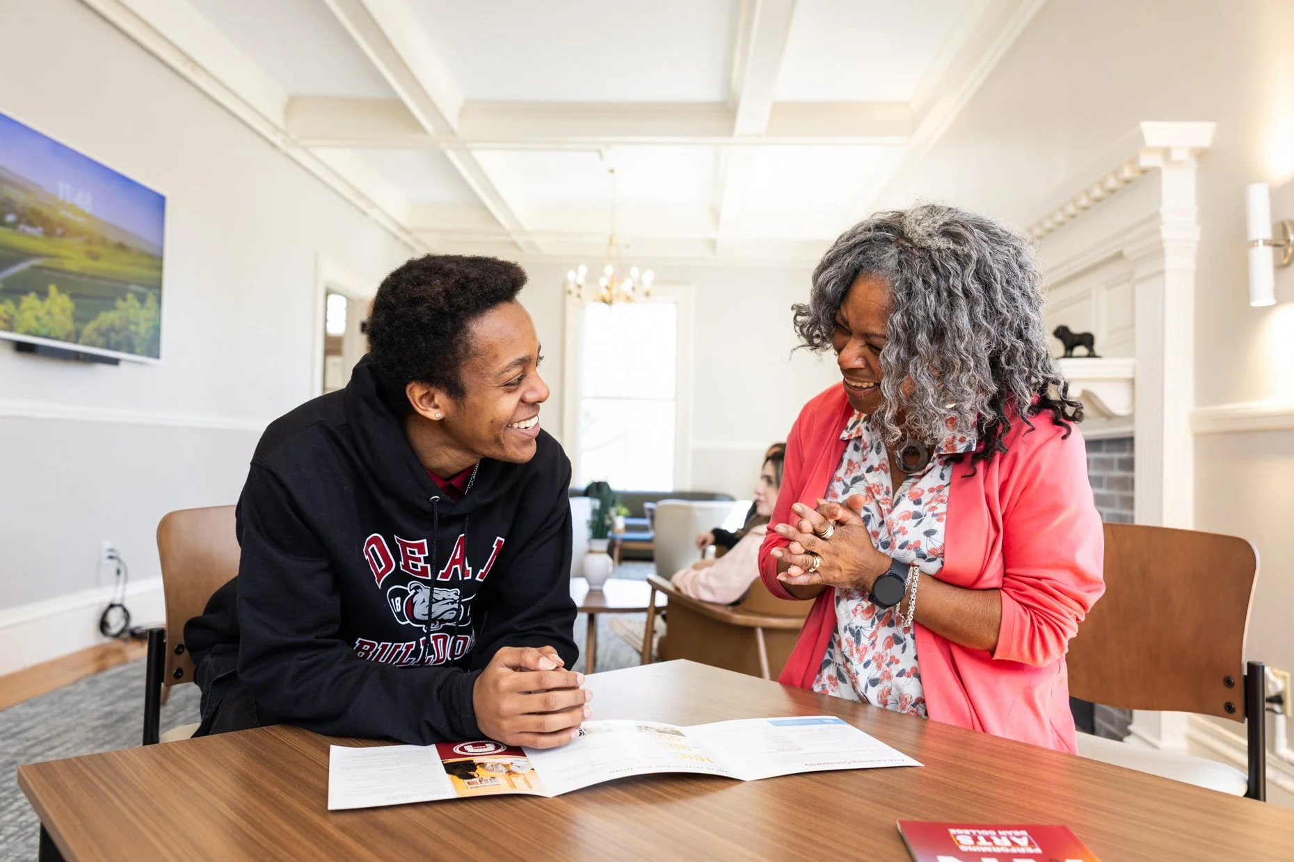 A young man and an older woman are smiling and engaging in conversation at a table in a bright, well-lit room. The young man is wearing a black hoodie with red and white lettering, and the woman is dressed in a pink blazer over a patterned blouse. Th