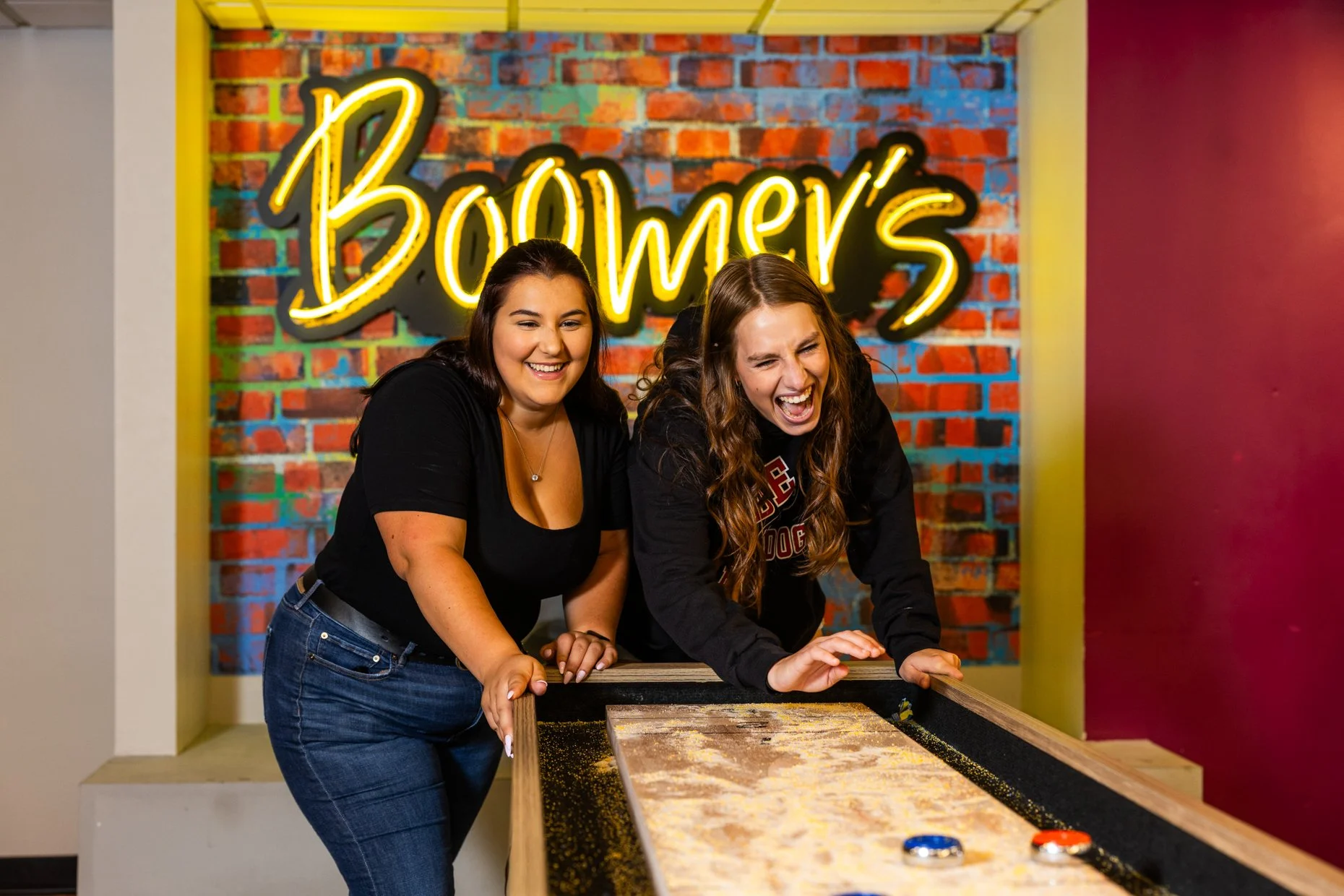 Two young women playing an arcade game at Bowler's arcade with a graffiti-style neon sign in the background.