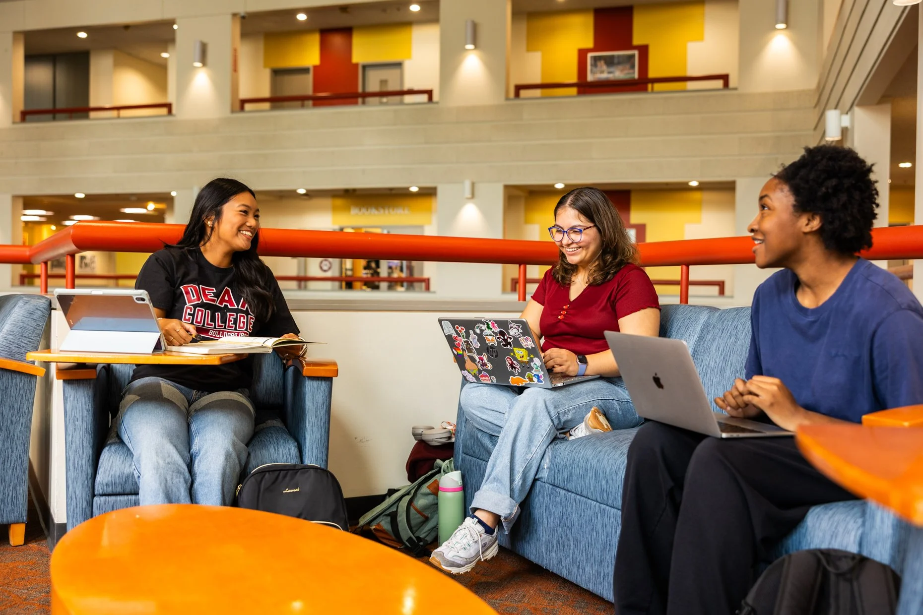 Three college students sitting on blue couches in a brightly lit indoor lounge, engaging in conversation and using laptops, with backpacks and water bottles on the floor.