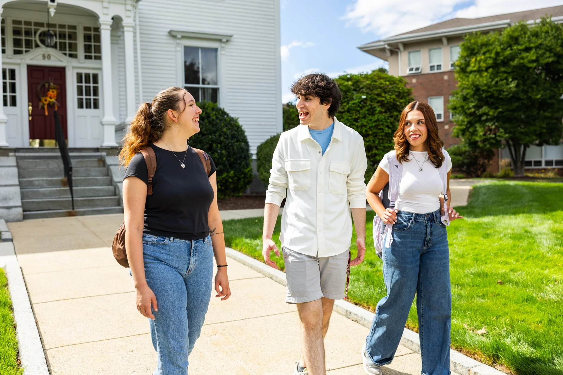 Three young people walking and talking on a sidewalk in front of houses on a sunny day, smiling and laughing.