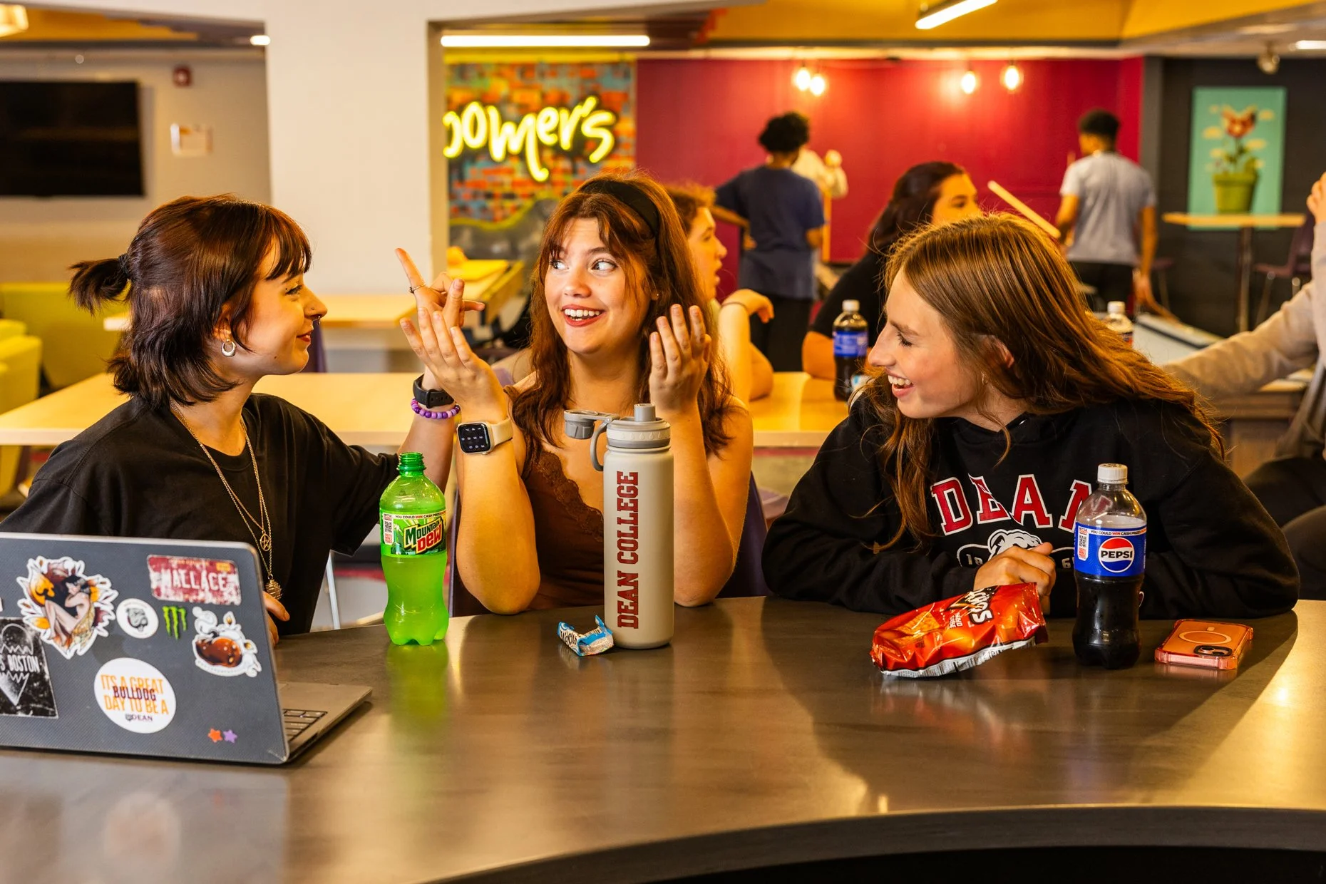 Three young women sitting at a table in a casual dining area, engaged in conversation and smiling. The table has drinks including Mountain Dew and Pepsi, snacks, and a laptop. The background features colorful walls and the neon sign 'Jowder's'.