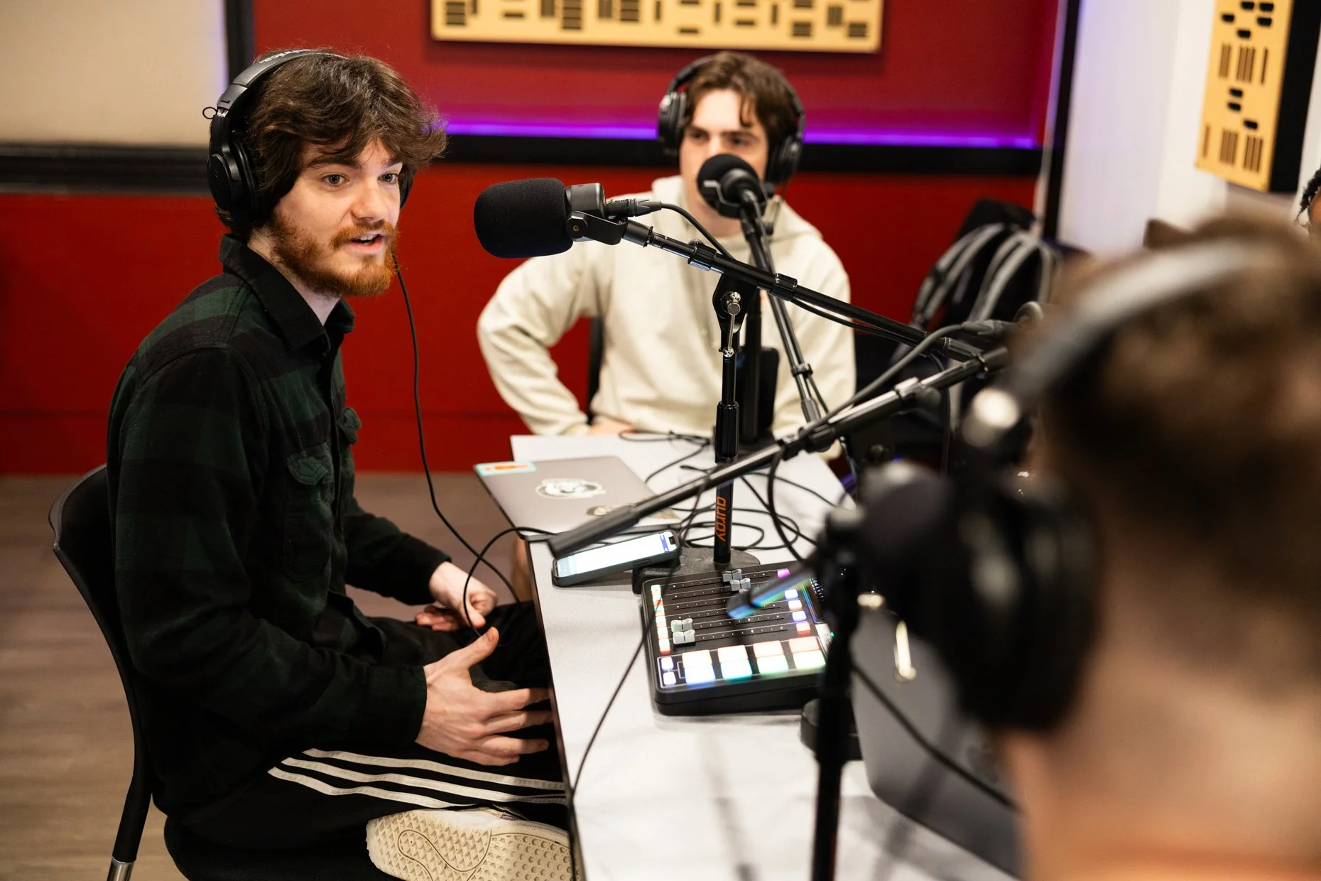 Two young men with headphones speaking into microphones in a recording studio. One is playing a keyboard and using a MIDI controller, the other is looking towards the microphones, with a red and purple background.