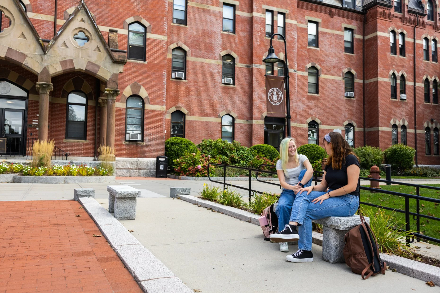 Two young women sit on a stone bench outside a brick college building, smiling and talking. One has blonde hair with green streaks, light skin, and is wearing a gray shirt. The other has brown hair, light skin, and is wearing a black shirt. They are 