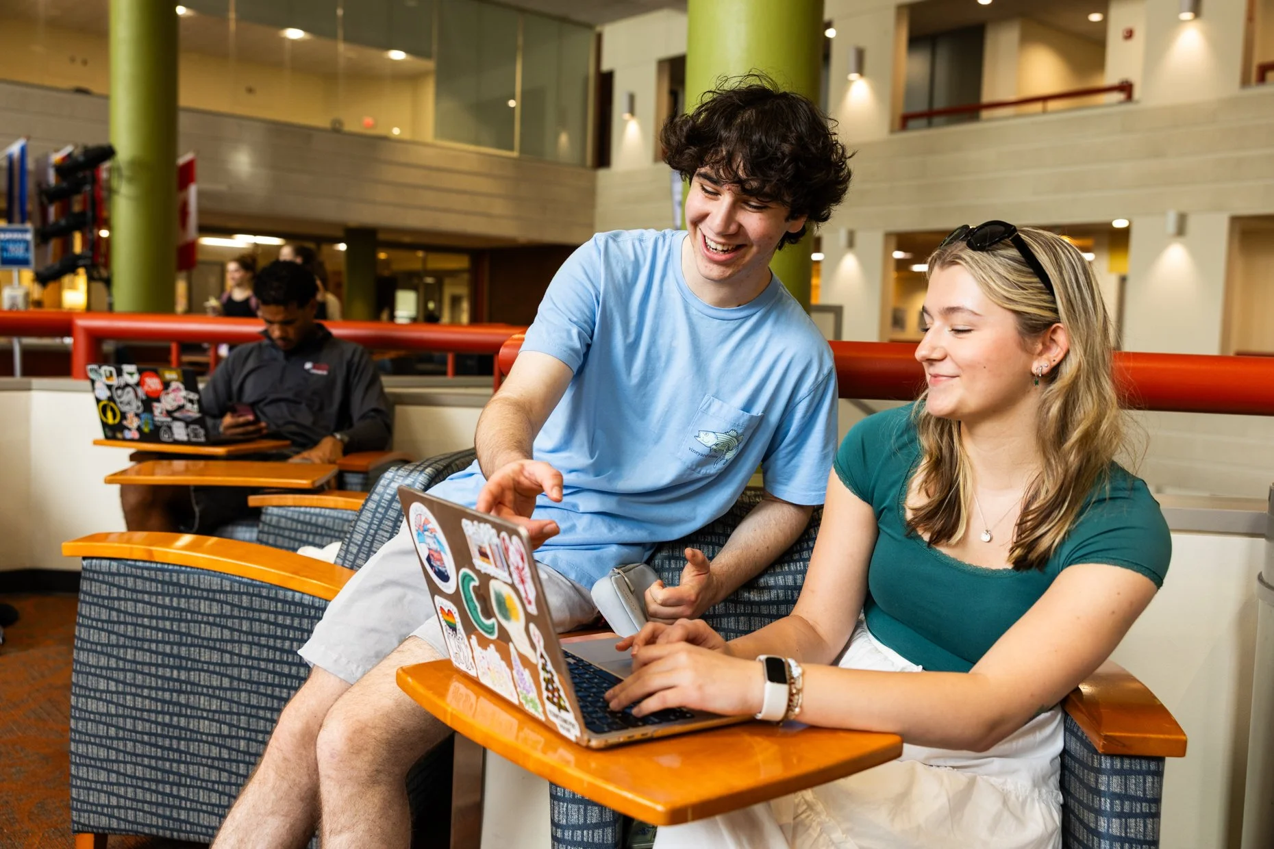 Two young adults sitting in a lounge area, smiling and looking at a laptop covered with colorful stickers. A man wearing a light blue t-shirt and a woman in a teal top with sunglasses on her head are engaged with the laptop, while a person in the bac