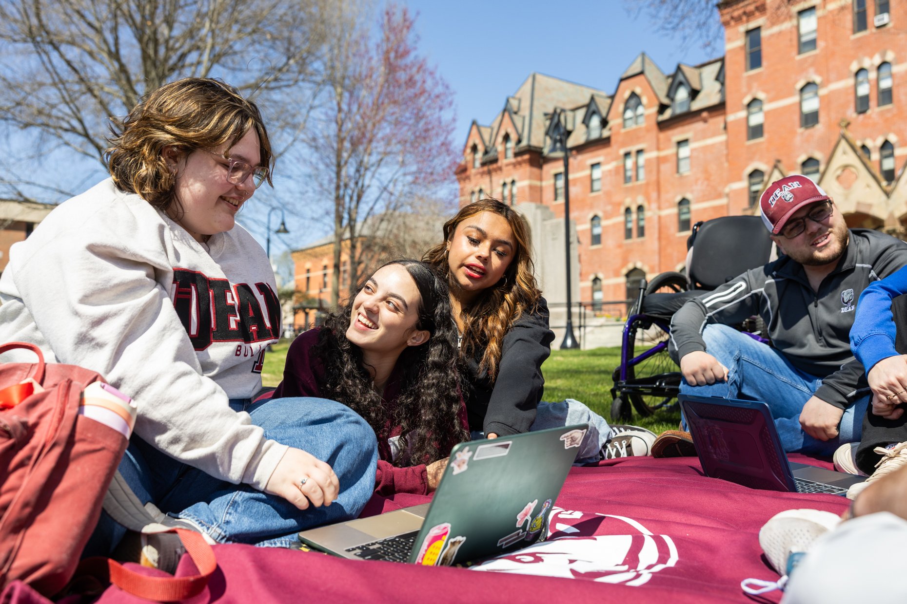 Group of four diverse young adults sitting outdoors on a red cloth, smiling and talking with laptops and backpacks around them, with a historic brick building and leafless trees in the background.