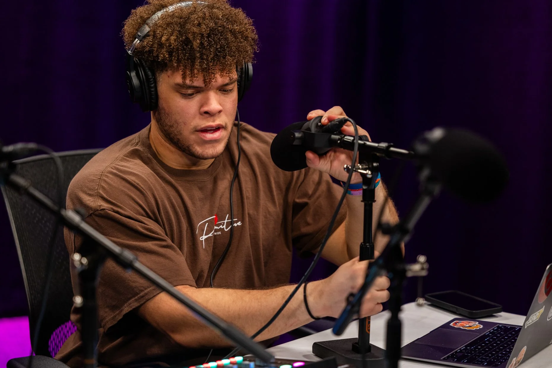 Young man with curly hair wearing headphones speaking into a microphone during a recording session, sitting at a table with a laptop and audio equipment.