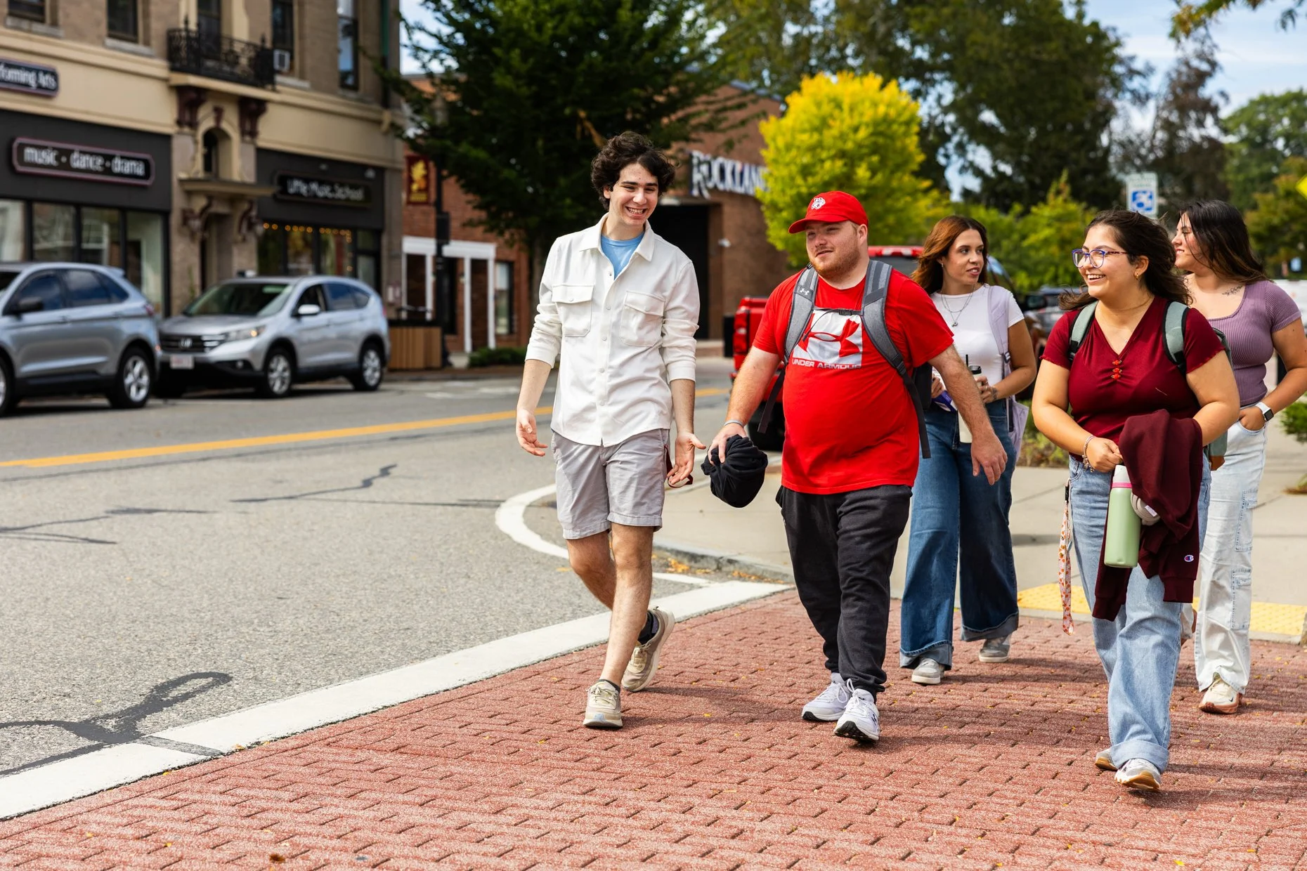 Six young people walking together on a city sidewalk, smiling and talking, with shops and cars in the background.