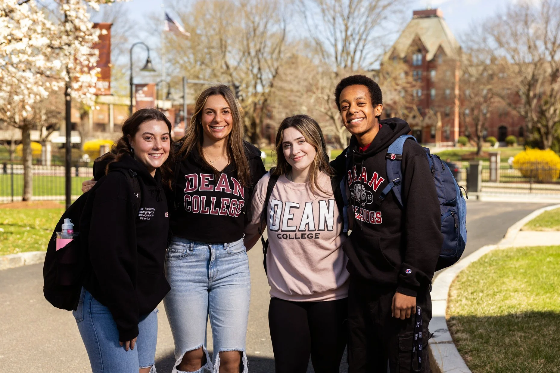 Four smiling students standing outside on a college campus with trees and buildings in the background. Three females and one male, all wearing casual clothes and backpacks, with some wearing college hoodies from Dean College.