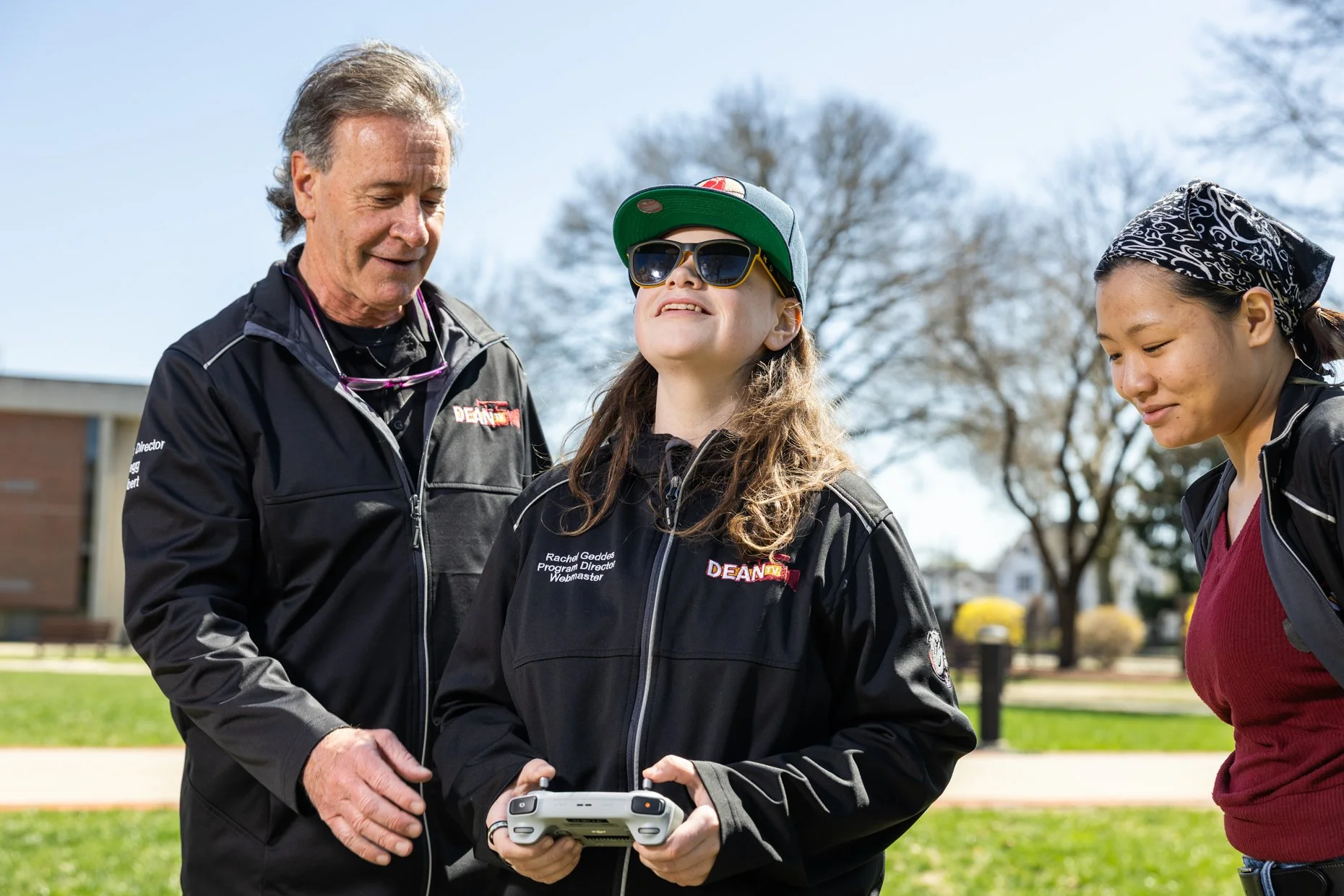 Three people outdoors, two women and one man, engaged with a drone controller. The man is smiling and wearing a black jacket, the woman in the center is wearing sunglasses, a baseball cap, and a black jacket with a logo, and the woman on the right is