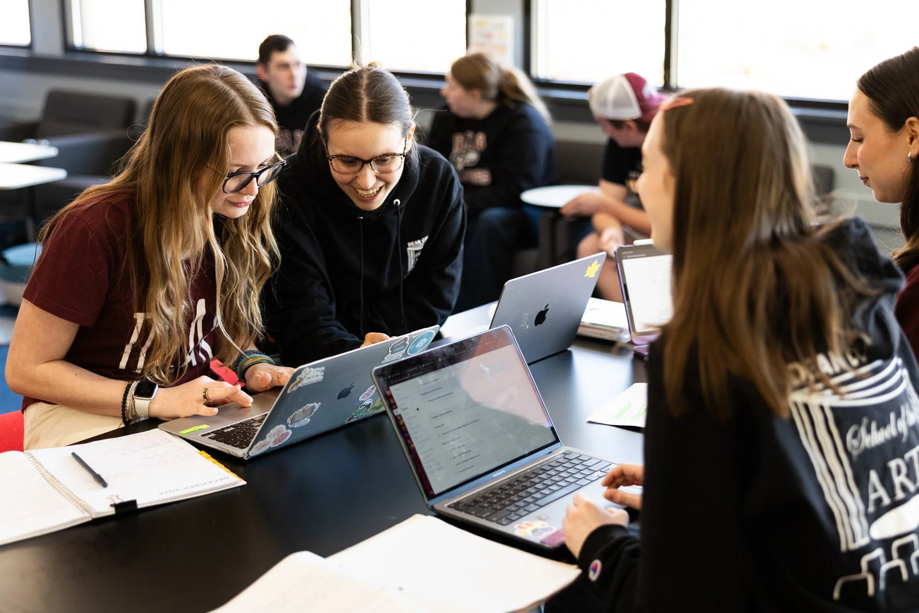 Group of young students sitting around a table with laptops and notebooks, engaged in a discussion or collaborative work in a classroom or study environment.