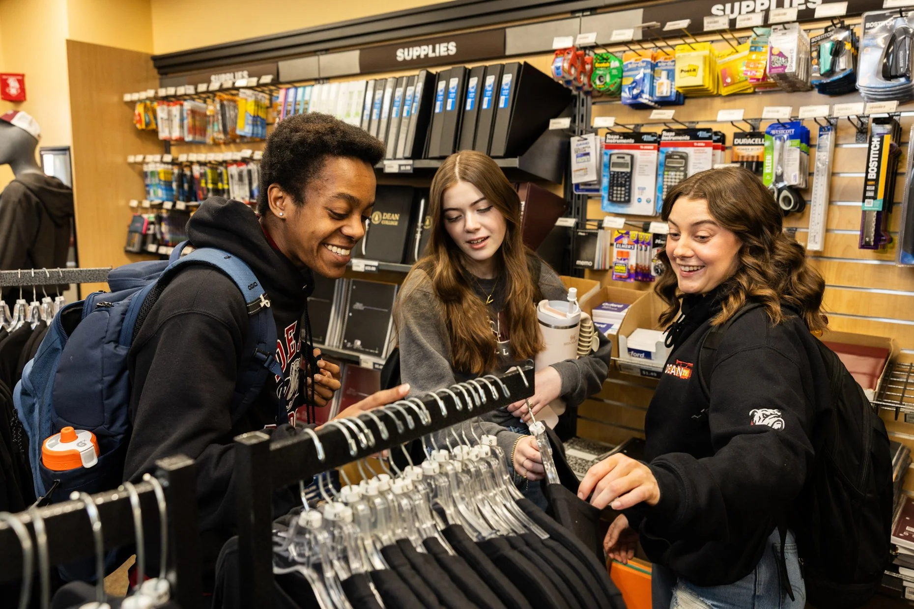Three young women shopping for clothes in a store aisle with shelves of supplies and notebooks in the background, smiling and chatting.
