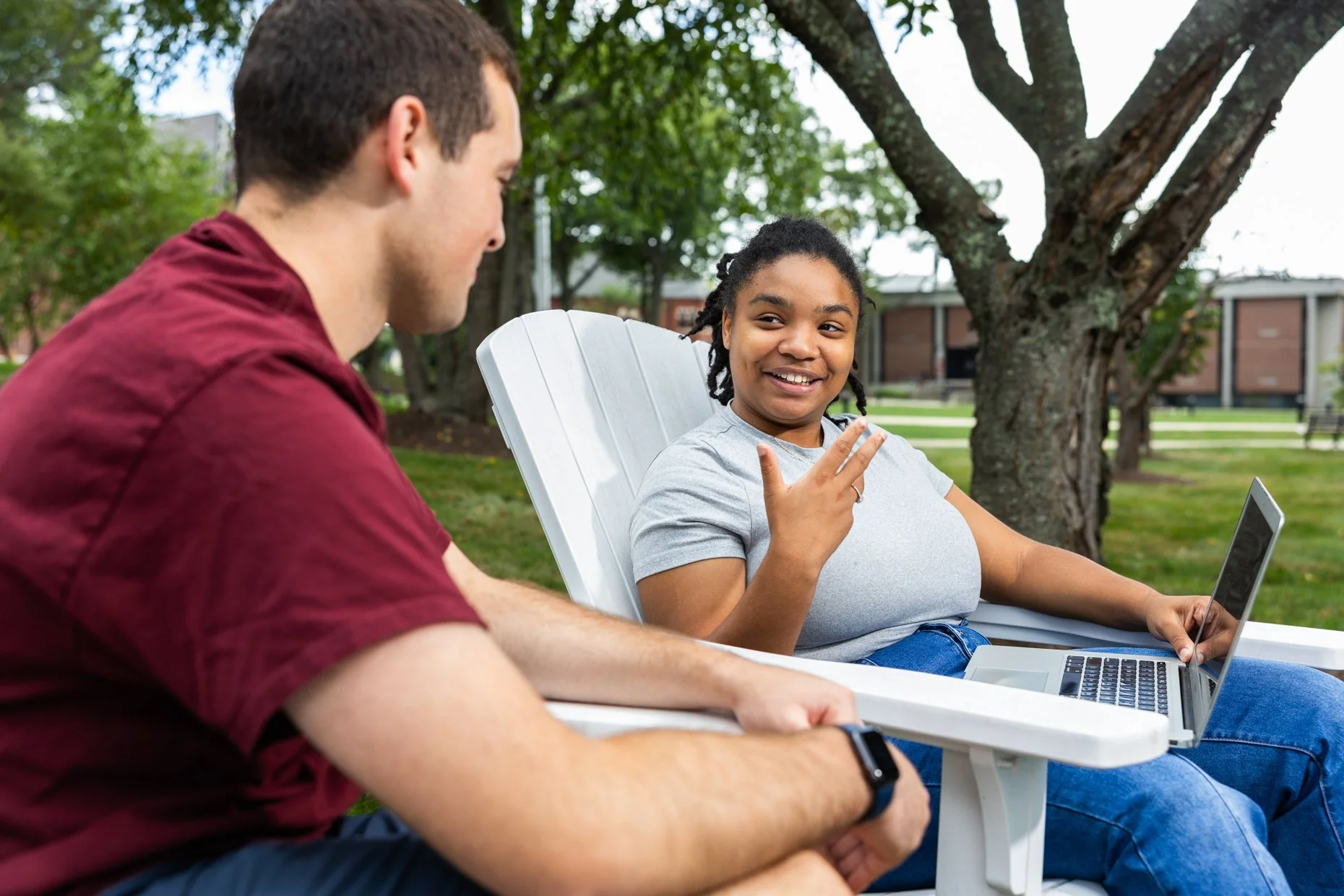 A woman with curly hair and a gray t-shirt sitting in a white outdoor chair, talking and making a peace sign with her fingers while looking at a man sitting across from her. The man is wearing a maroon shirt, has short brown hair, and is wearing a sm