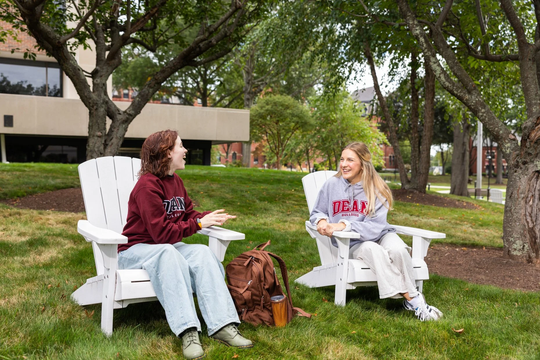 Two young women are sitting on white outdoor chairs in a park, engaged in conversation and smiling. One wears a maroon sweatshirt and light jeans, while the other wears a gray hoodie with university branding and striped pants. They are surrounded by 