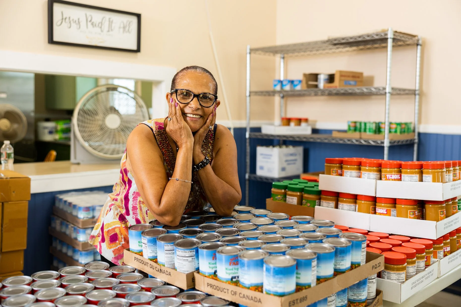 A woman with glasses and a colorful sleeveless top smiling and resting her face in her hands behind a display of canned goods and jars of peanut butter in a food pantry.