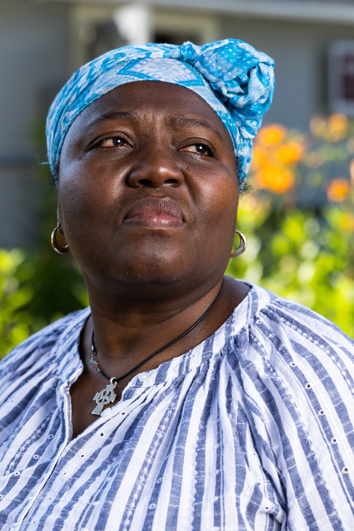 A woman with dark skin wearing a blue patterned headscarf and a striped shirt, outdoors with blurred yellow flowers in the background.