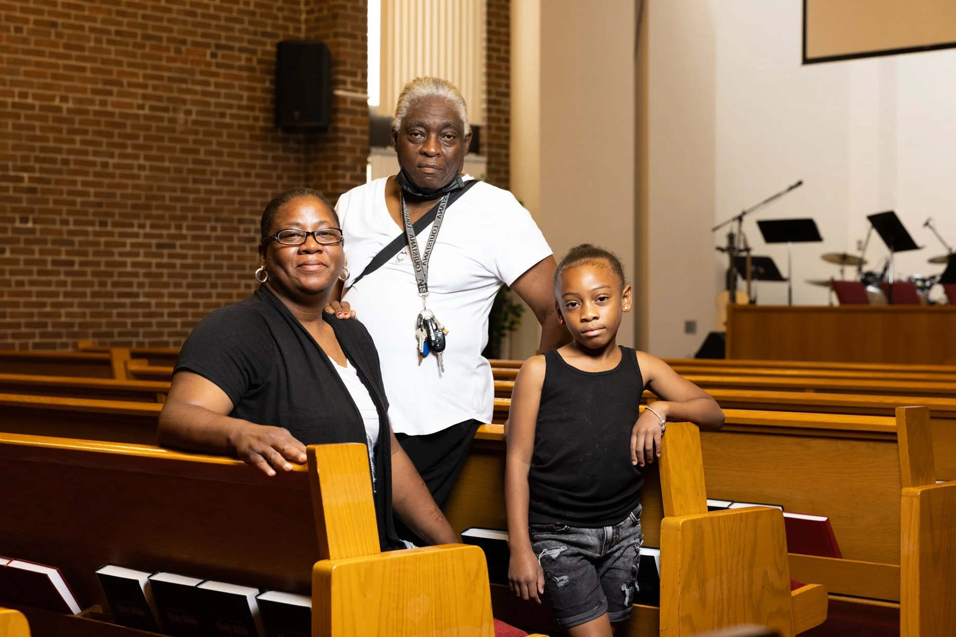 Three people, two women and a young girl, inside a church with wooden pews and a brick wall. The woman on the left wears glasses and a black top, the man in the middle has gray hair, a lanyard with keys, and a white shirt, and the girl on the right w