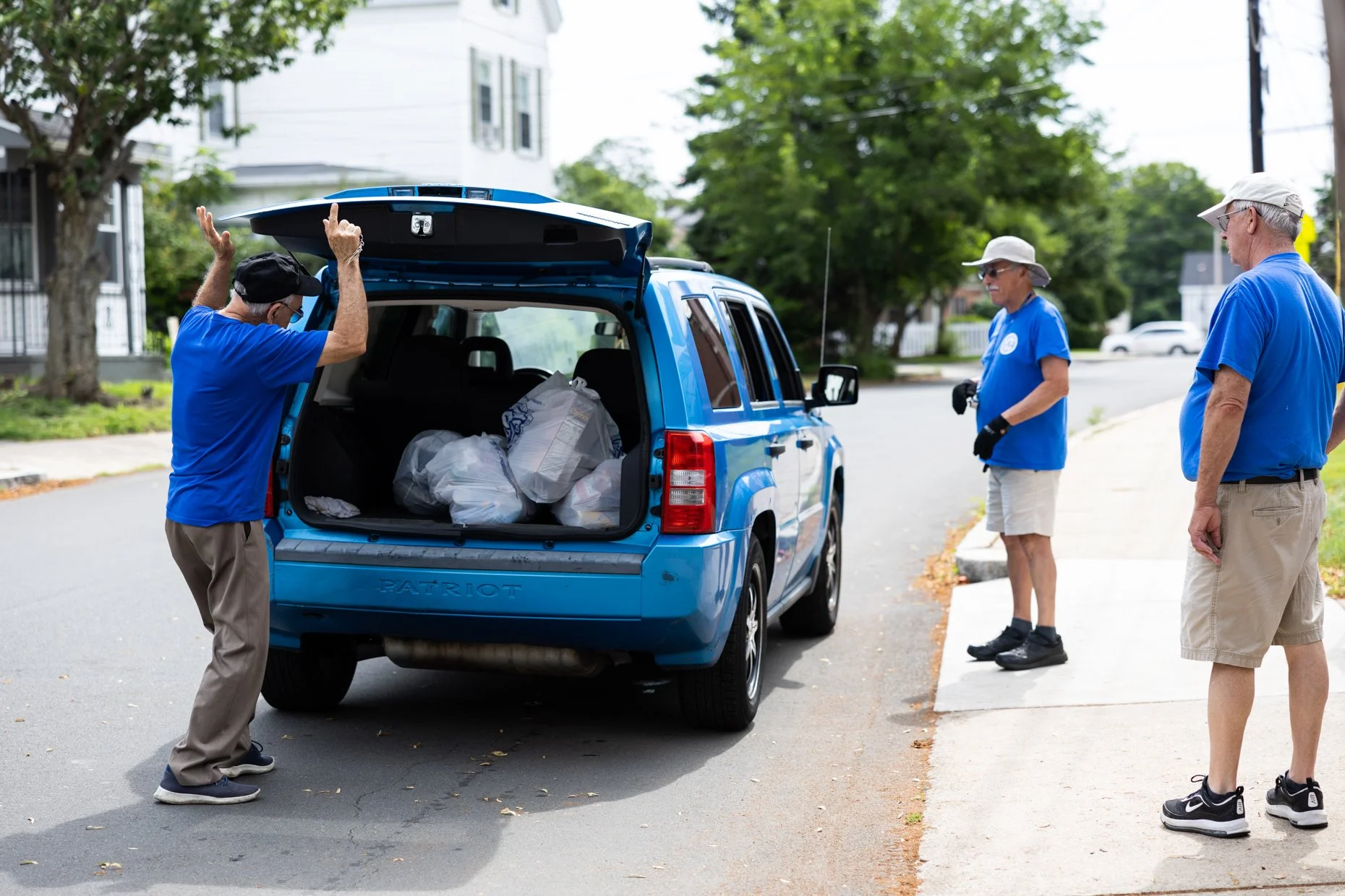Three elderly men in blue T-shirts and beige shorts or pants standing on a sidewalk, loading bags into the back of a blue SUV.