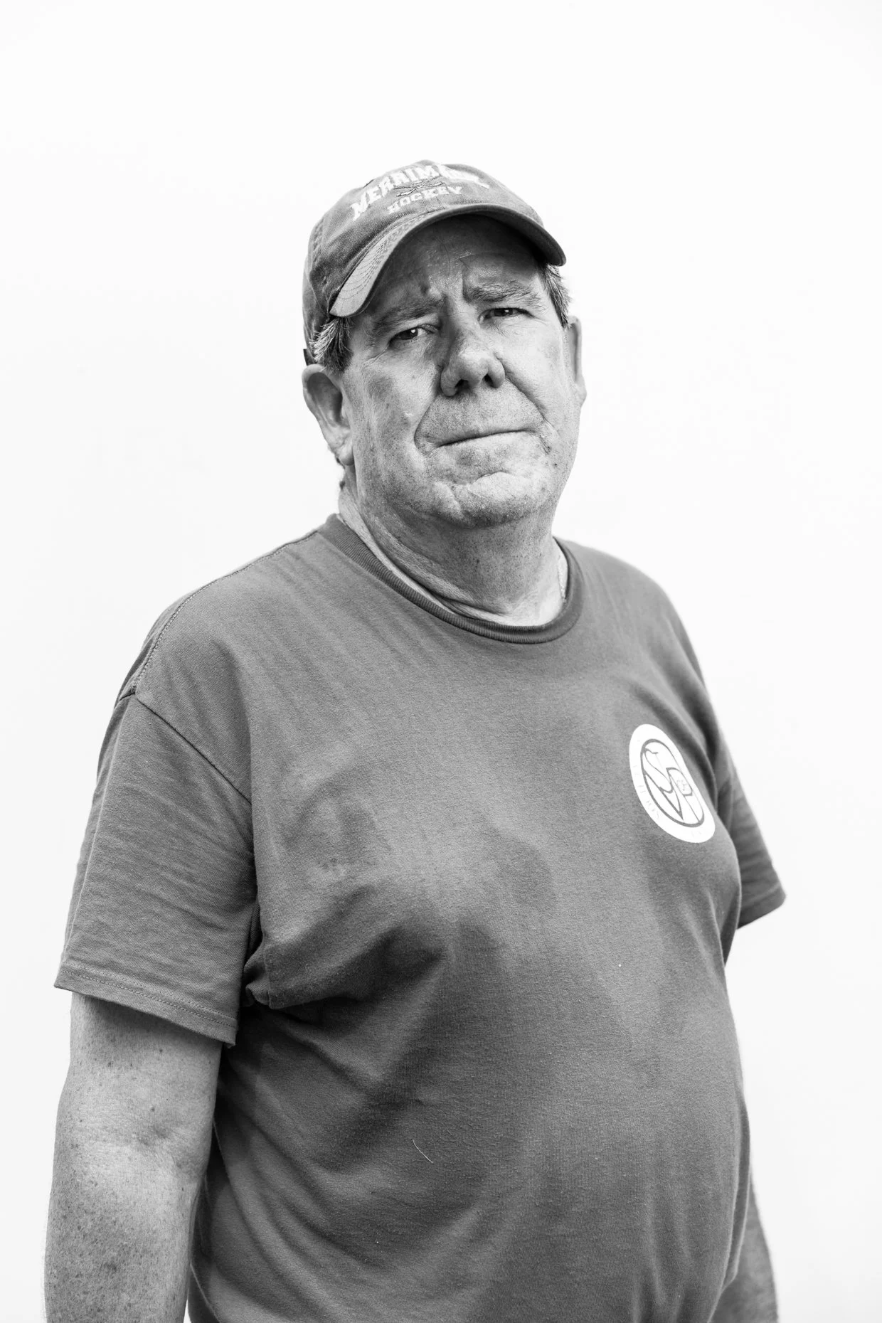 Black and white photo of an older man wearing a cap and a t-shirt, standing in front of a plain white background.