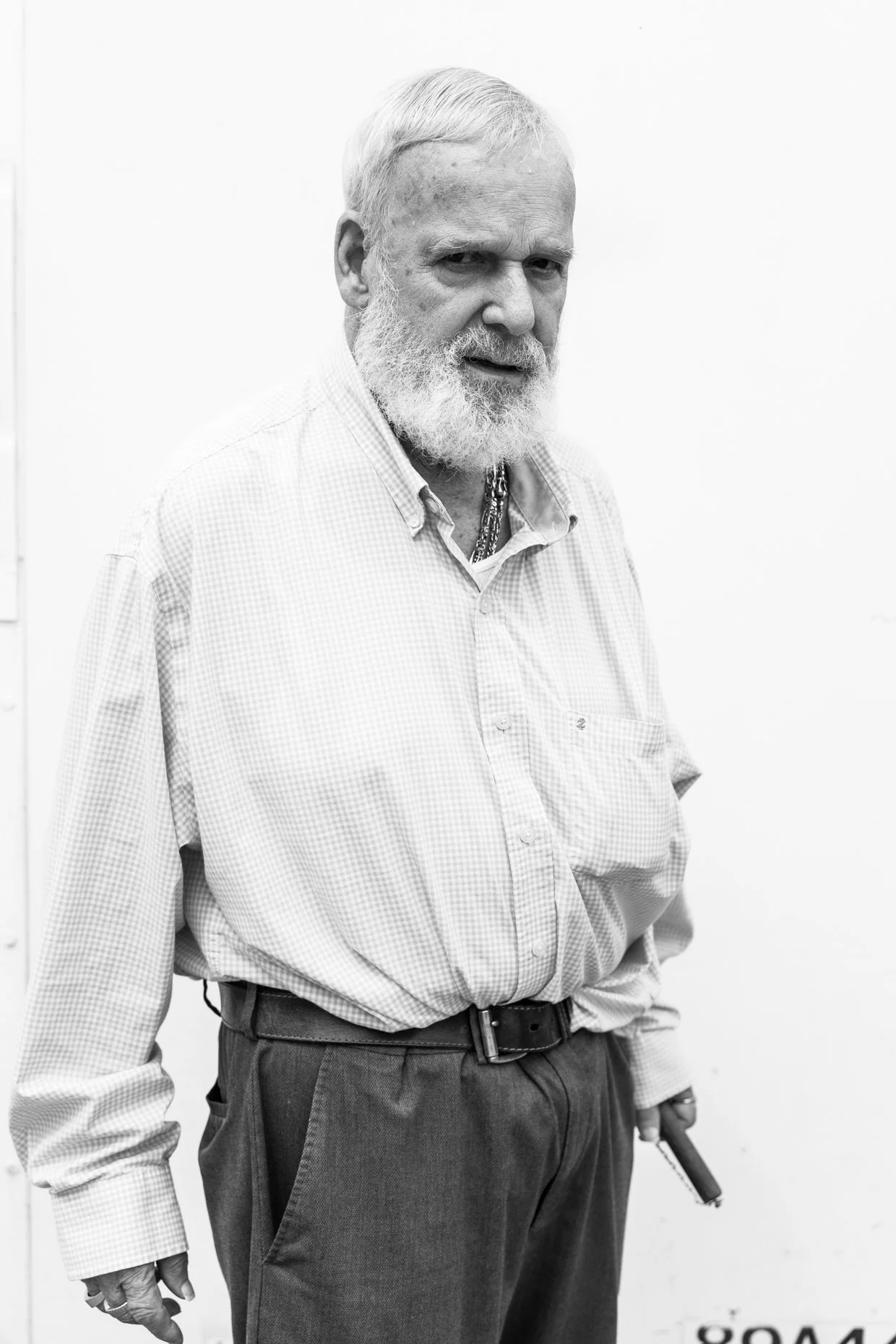An elderly man with a white beard and hair, wearing a light-colored checkered shirt and dark trousers, holding a gun in his left hand, standing against a plain white background.