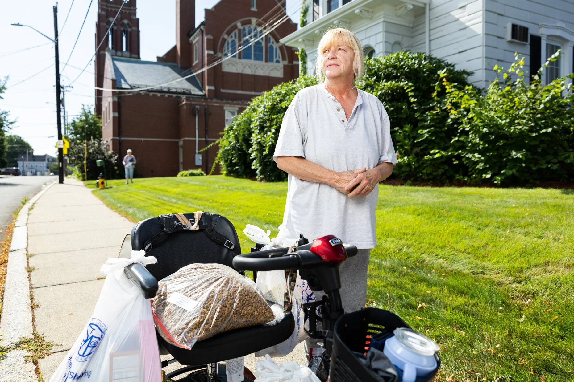 An elderly woman with blonde hair stands outside on a sunny day near a bicycle loaded with bags, groceries, and other items. Behind her are a green lawn, large bushes, and a white house. In the background, there is a church with brick walls and a sma