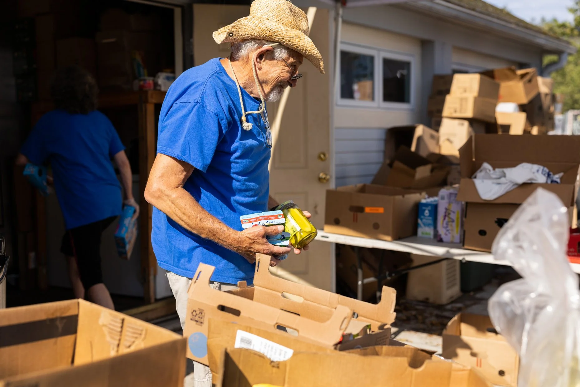 Elderly man with gray hair, beard, glasses, wearing a blue shirt and straw hat, sorting through food donations in a garage filled with boxes and shelves, with a woman in blue in the background.