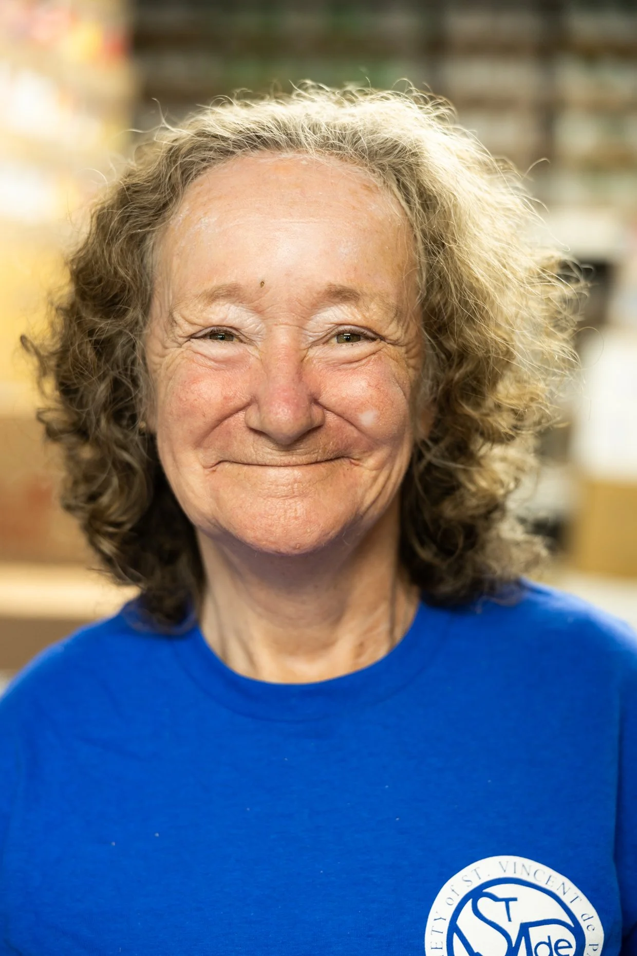 A smiling elderly woman with curly gray hair wearing a blue shirt with a logo.