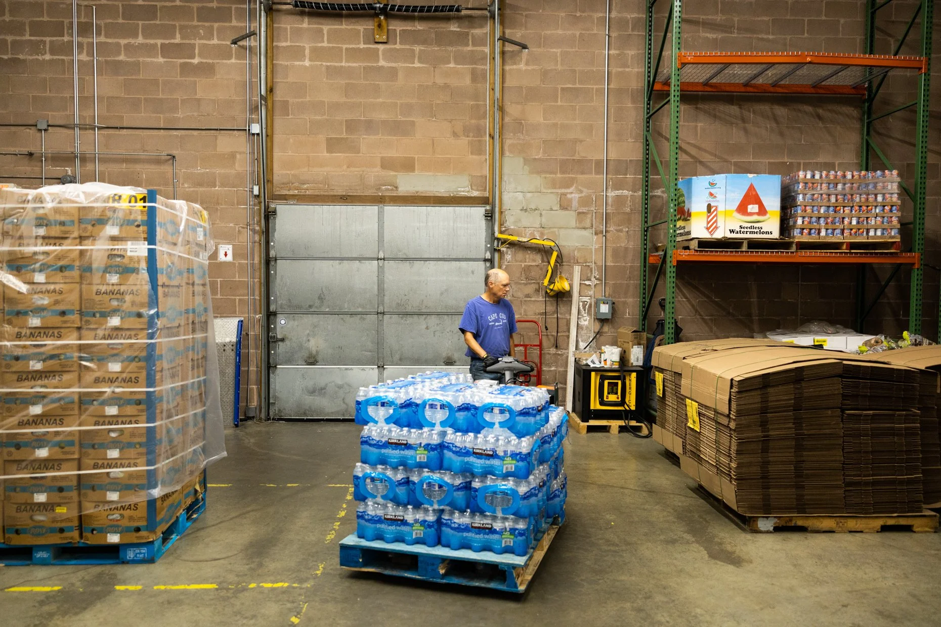 Inside a warehouse with a man standing near bottled water and stacks of cardboard boxes, pallets of bananas to the left, and shelving units with various items to the right, brick walls and a large metal door in the background.