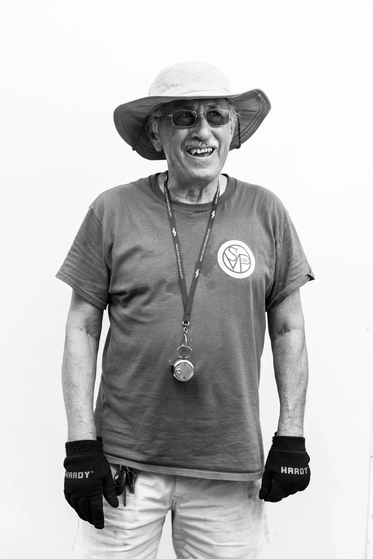 An elderly man wearing a sunhat, sunglasses, a T-shirt with a logo, gloves, and a whistle around his neck, smiling and standing against a plain background.