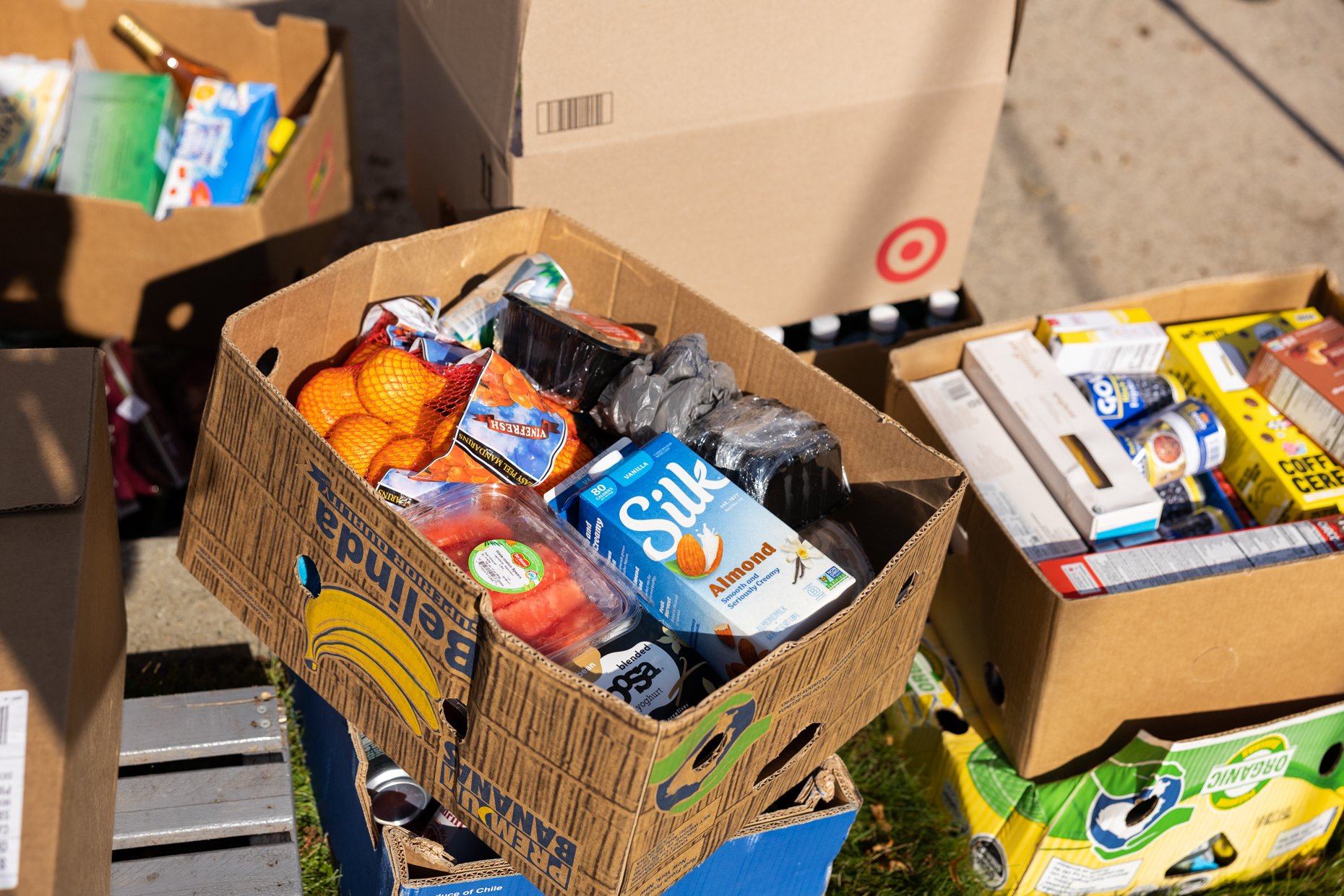 Several cardboard boxes filled with various groceries, such as juice, snacks, and canned goods, are stacked outdoors.