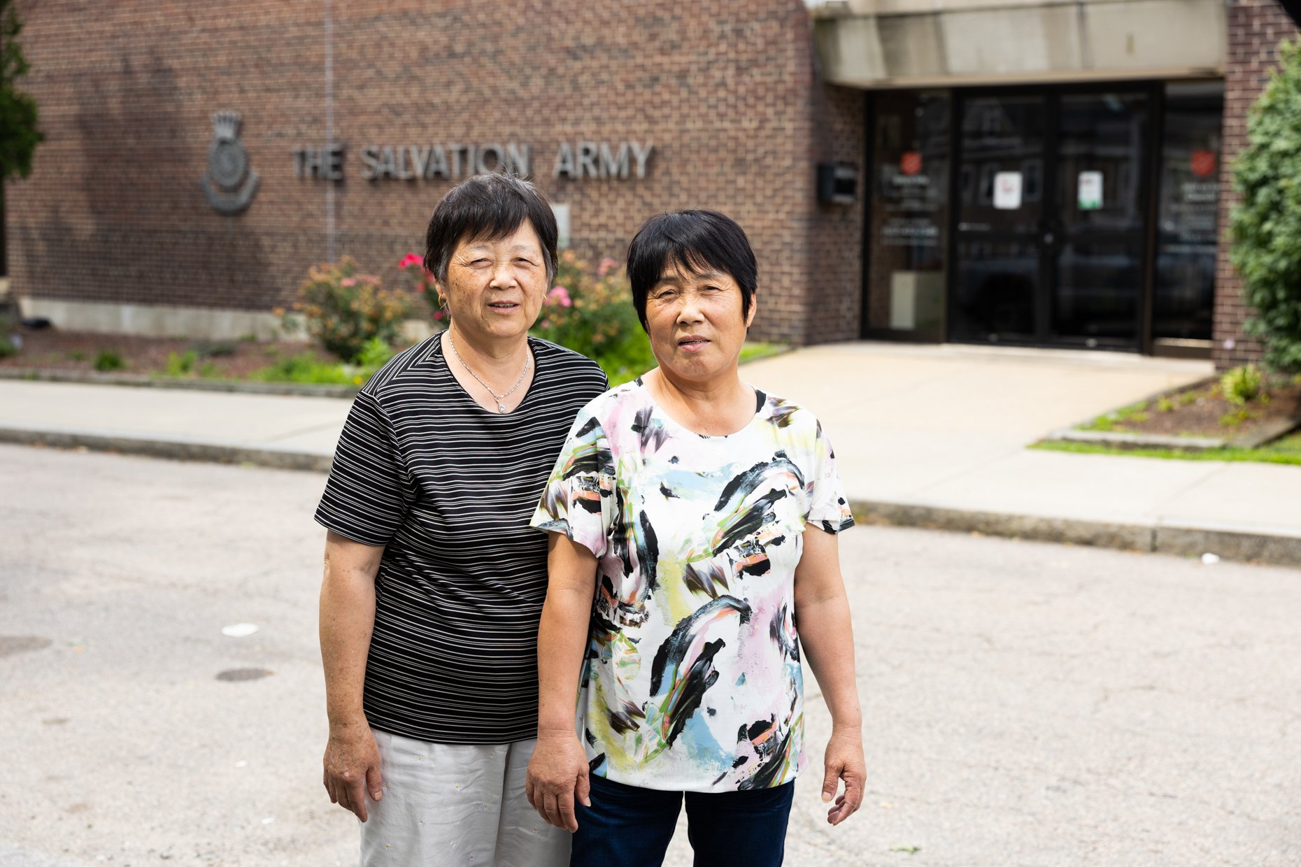 Two elderly women standing outside a brick building with a sign that reads "The Salvation Army." They are standing on a sidewalk, with greenery and flowers in the background.