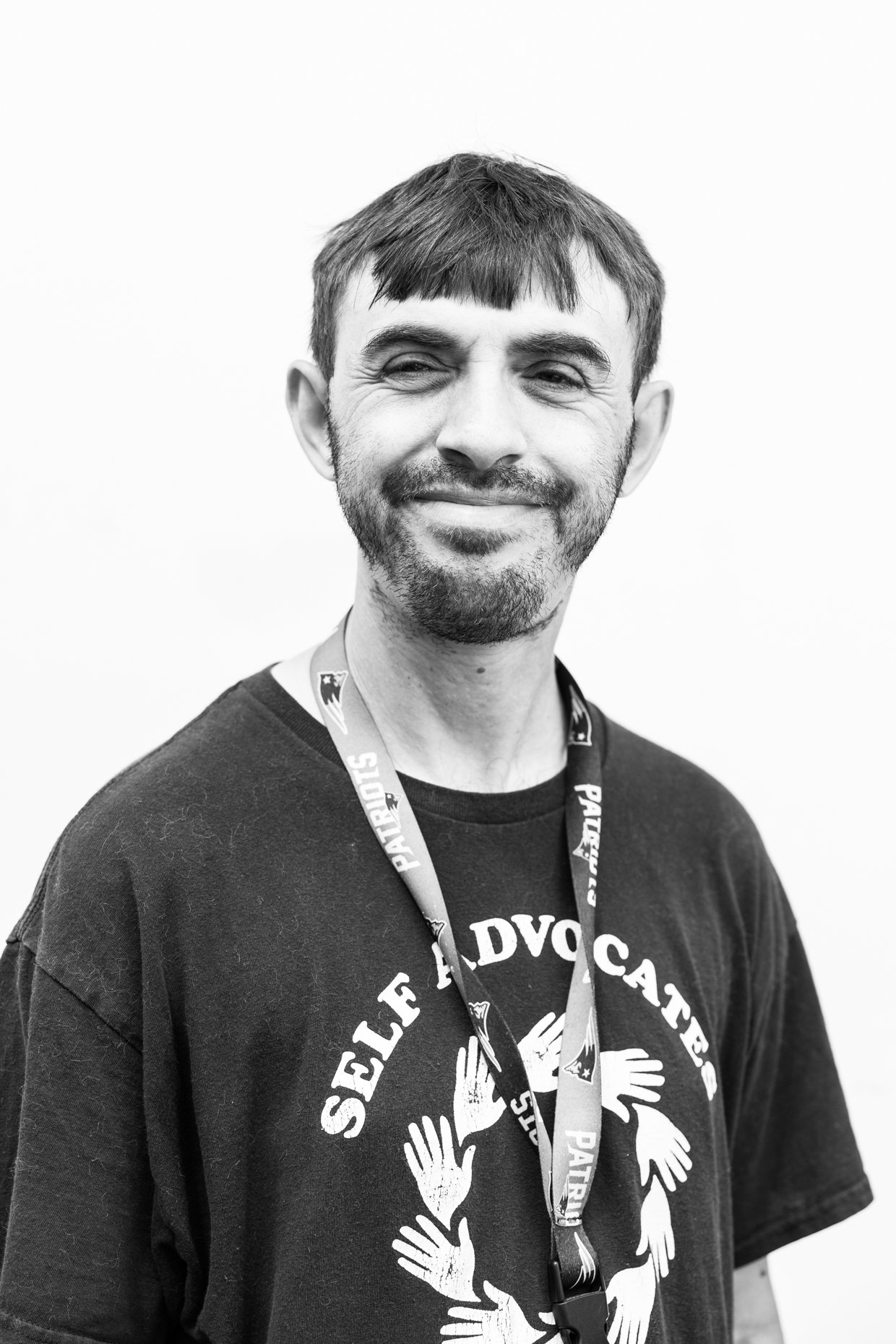 Black and white photo of a man with a friendly smile, wearing a lanyard and a T-shirt that has a circular slogan with several hand illustrations around it.