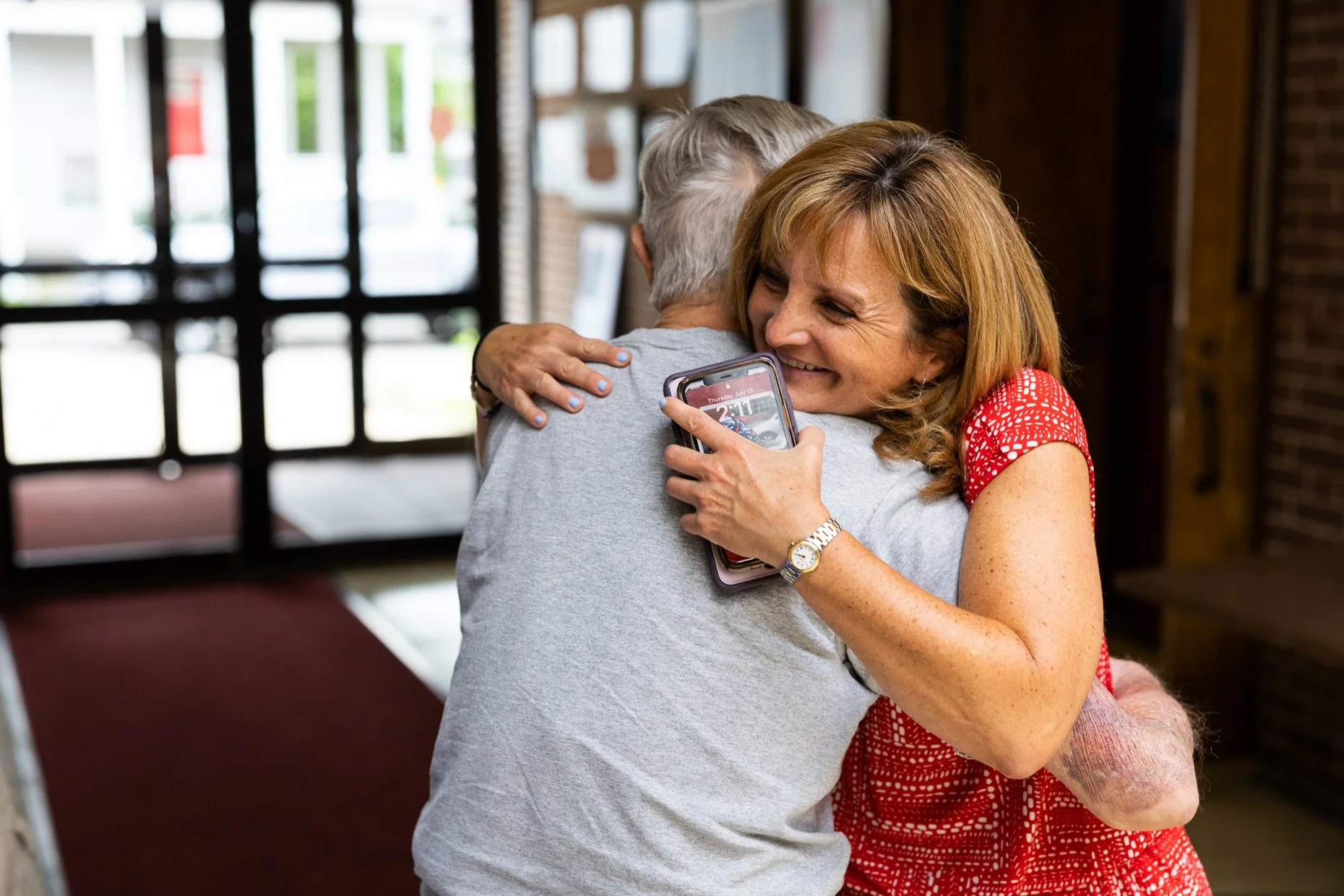 A woman with red hair hugging an elderly person with gray hair inside a building, smiling and holding a smartphone with a picture showing on the screen.