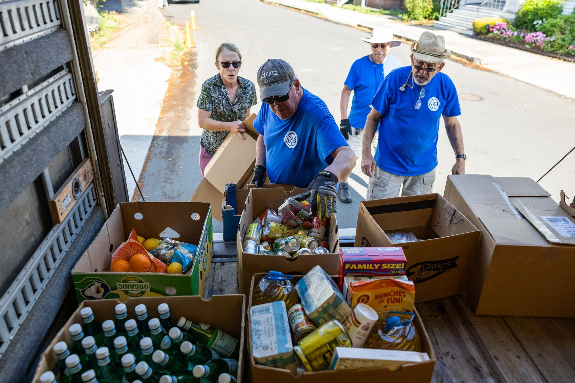 Group of people organizing donated food items in a truck bed, including canned goods, bottled drinks, and boxes, on a residential street with houses and flowers in the background.