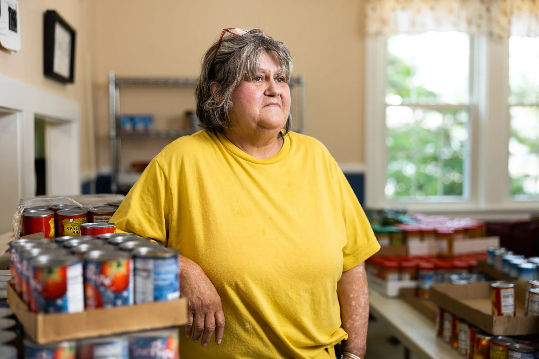 A woman with gray hair and glasses on her head, wearing a yellow shirt, standing in a room with windows and shelves of food items, looking serious or contemplative.