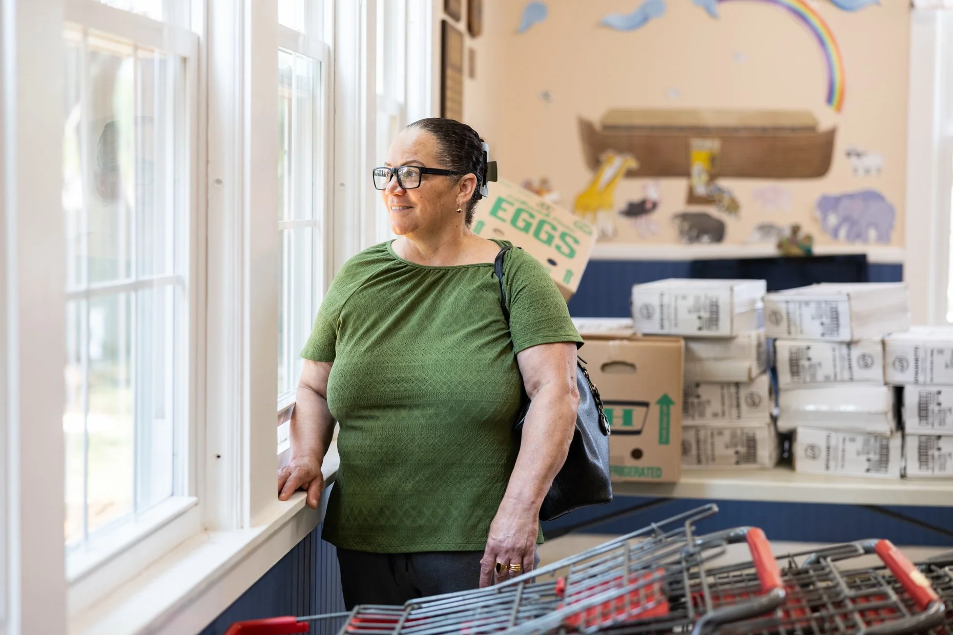 Woman in green shirt with glasses looking out a store window surrounded by boxes and shopping carts.
