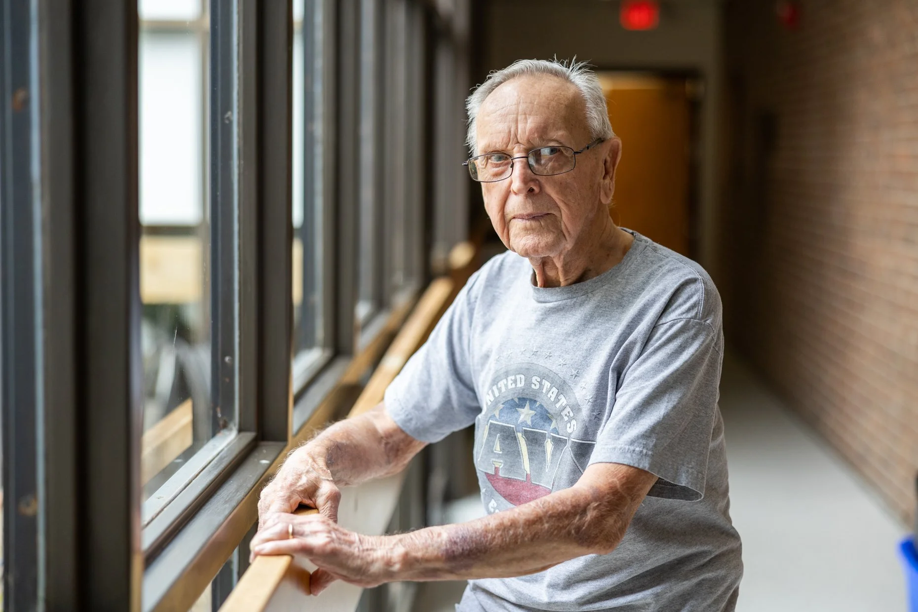 An elderly man with glasses standing by a window, looking outside with a serious expression. He is wearing a gray t-shirt with a United States logo and appears to be holding a wooden railing or ledge with his hands.