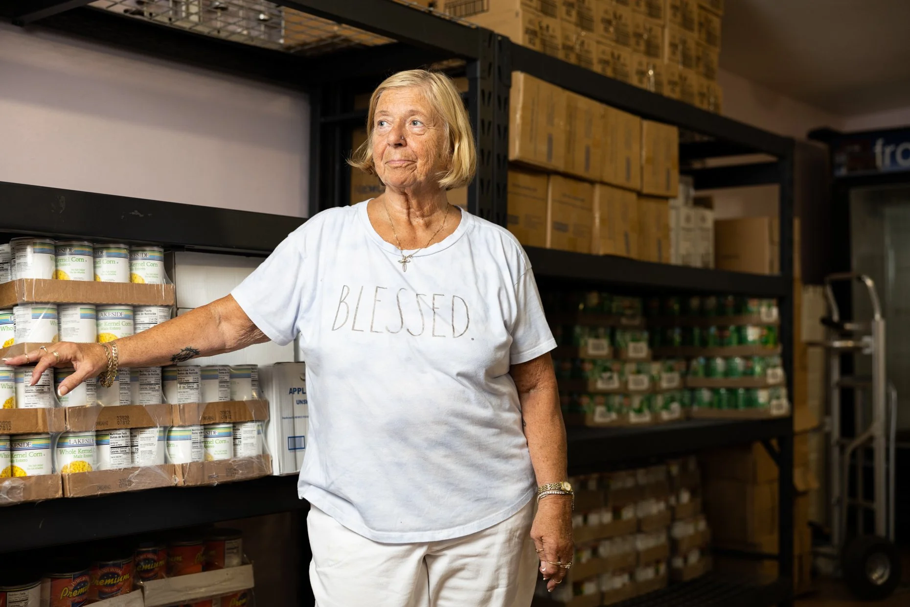 An elderly woman with blonde hair, wearing a white t-shirt with 'BLESSED' written on it, standing in a storage room with shelves filled with canned goods and boxes, reaching out to touch a can of whole kernel corn.
