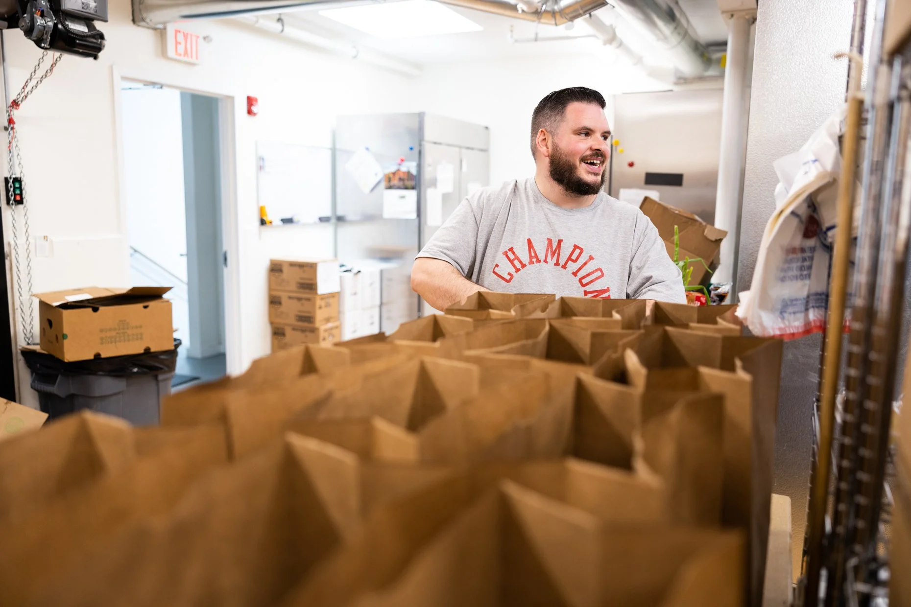 A man in a gray t-shirt with 'Champion' written on it handles paper bags in a room filled with boxes and supplies, seemingly organizing or preparing items.
