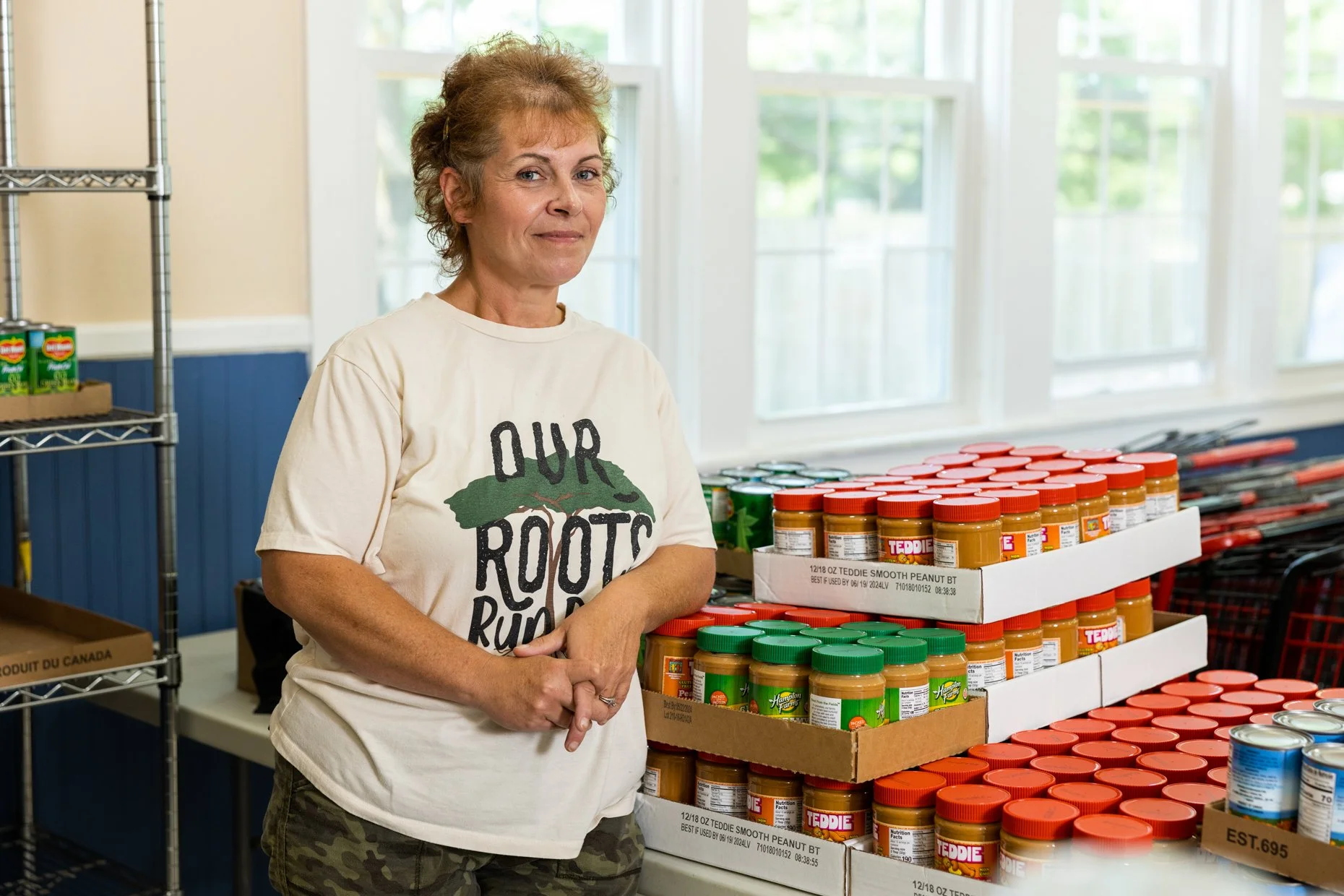 A woman standing in a room with large windows, behind a table with peanut butter jars stacked in boxes. She is wearing a white t-shirt with a green tree and the text 'OUR ROOTS RUN DEEP' and camouflage pants, with curly short hair, and is looking at 
