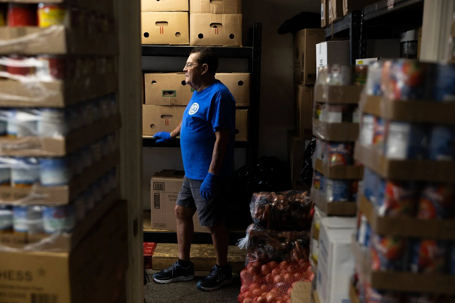 A man in a blue t-shirt, black shorts, and black sneakers stands in a storage room or pantry, surrounded by shelves filled with boxes of food. He is wearing blue gloves and appears to be organizing or preparing food items, with onions in a mesh bag o