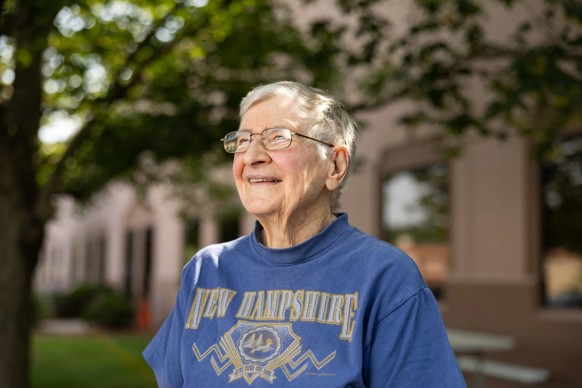 Smiling elderly man with glasses standing outdoors with trees and a building in the background, wearing a blue shirt with "New Hampshire" written on it.