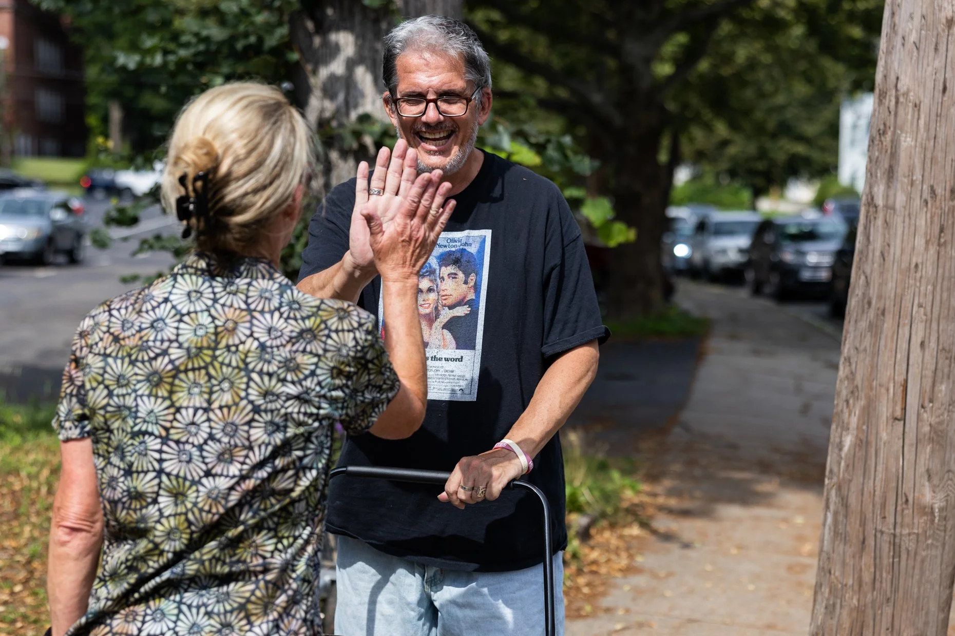 An elderly woman and a middle-aged man exchange a high five outdoors on a sidewalk, with trees, parked cars, and a building visible in the background.