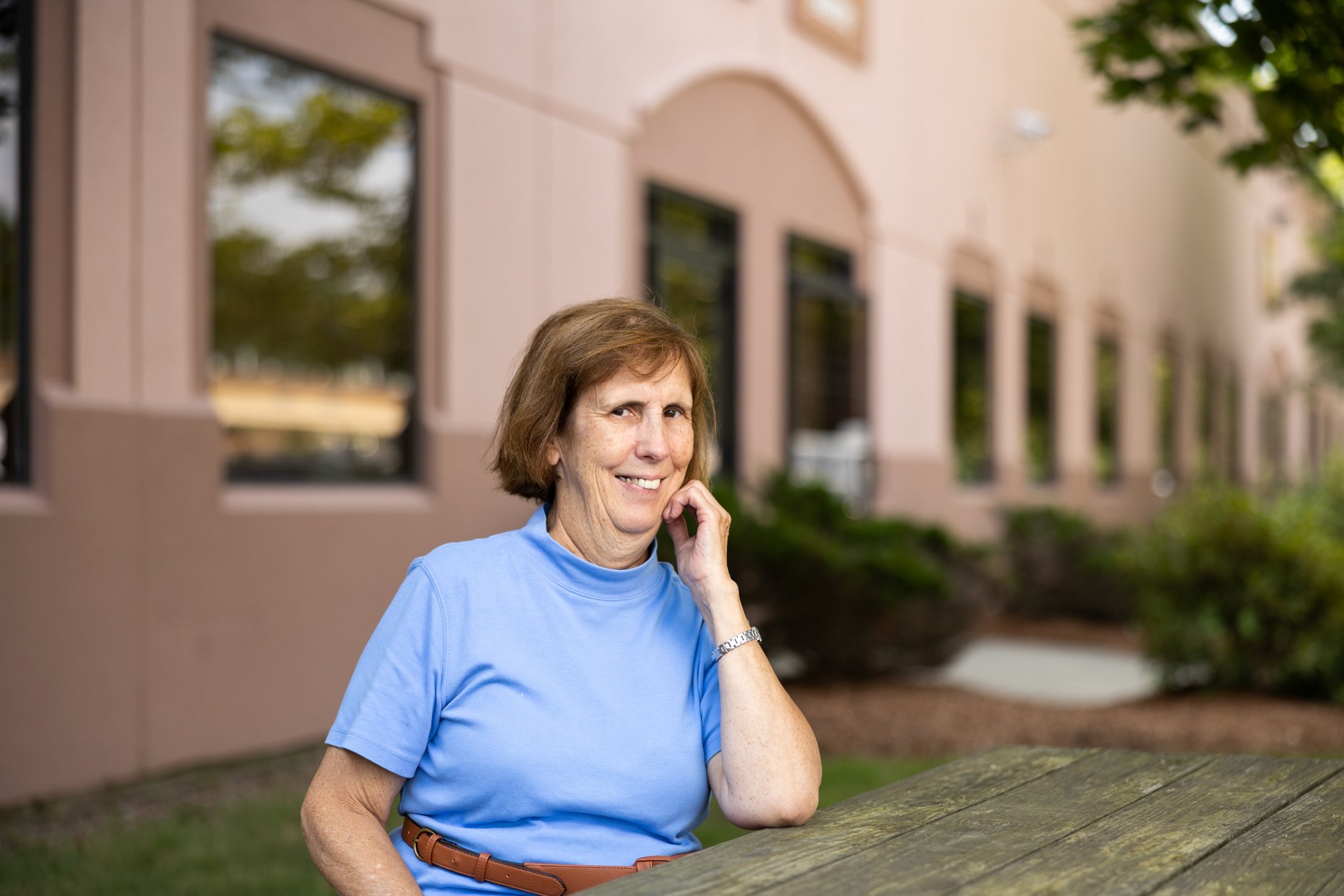 A smiling older woman with short brown hair, wearing a light blue T-shirt, standing outdoors near a wooden table in front of a beige building with multiple windows, with trees and greenery in the background.